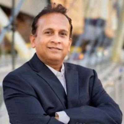 A man with a beard and mustache, wearing a dark blazer and black turtleneck, looking directly at the camera with a serious expression, blurred lights in the background.