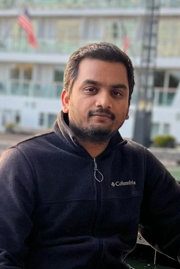 A man with dark hair and facial hair wearing a dark Columbia jacket, sitting outdoors with a blurred modern building in the background.