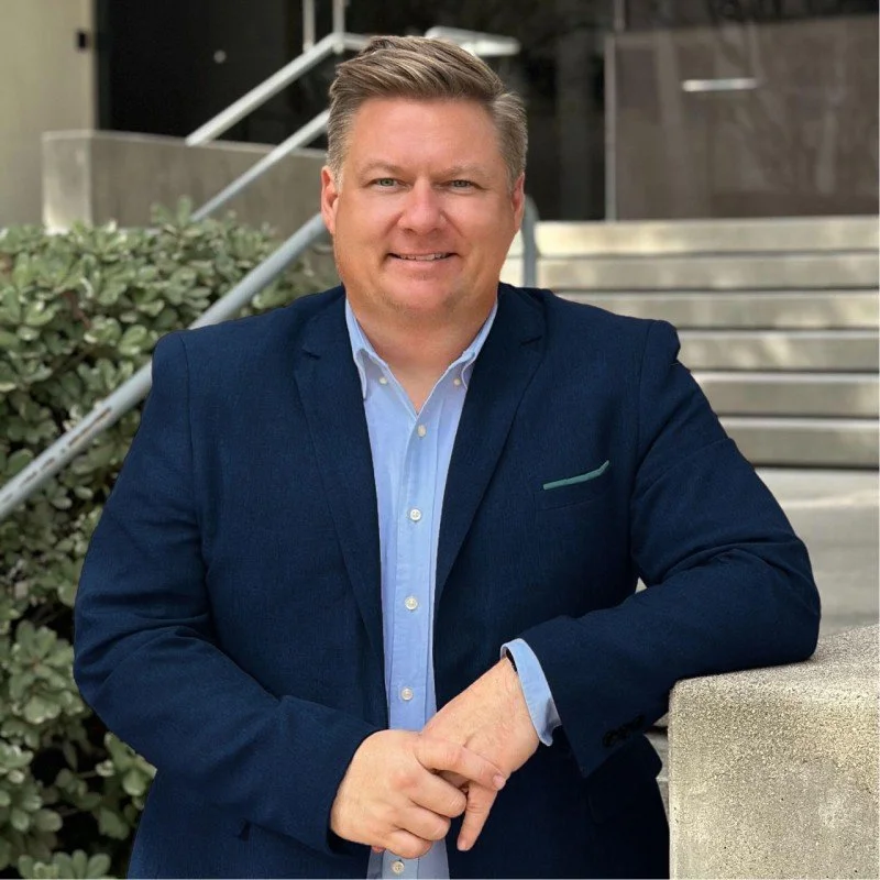 A man in a red blazer and white shirt smiling at the camera with a neutral background.