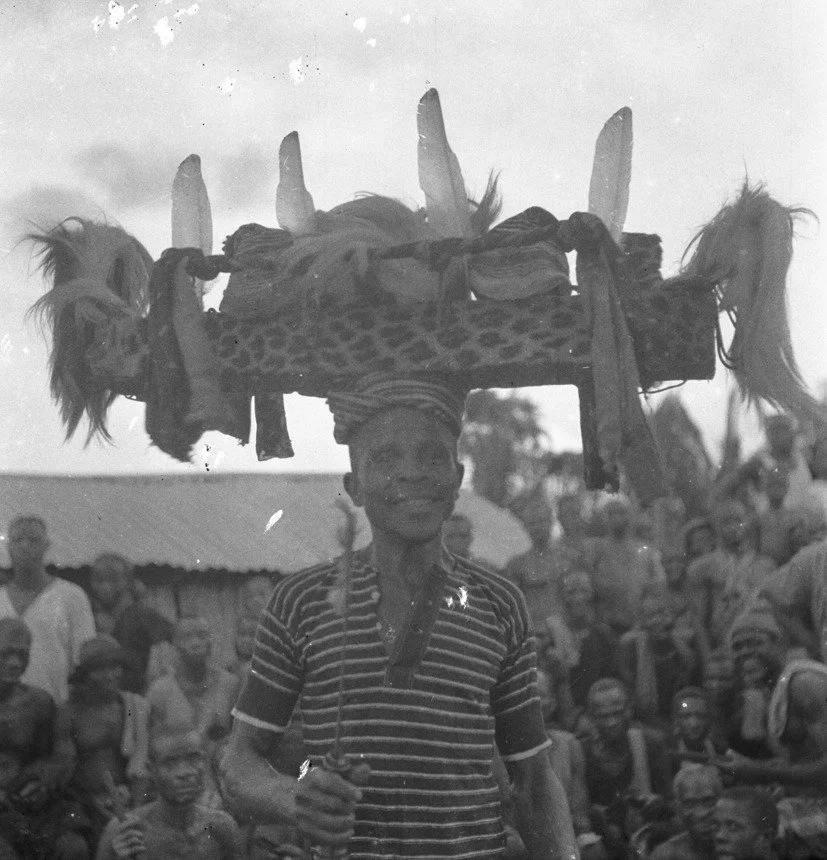 [Igbo] Ohafia war dance: The Ohafia war dance iri agha. Adult male carrying a headhunters trophy (oyaya) on his head. The trophy is a rectangular shaped wooden frame decorated with feathers, leopard fur, cloths and animal fur at either end. He is wea