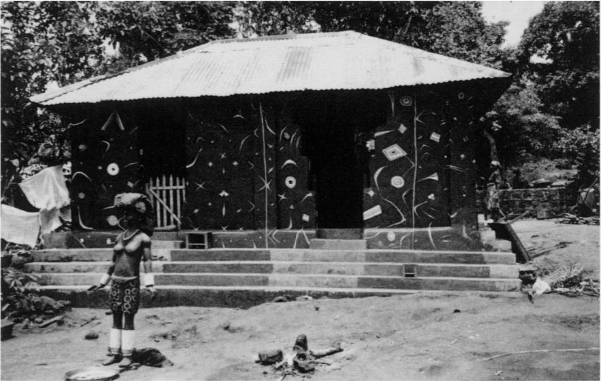 'Shrine to the Goddess Eke at Uke Market. Newly repainted with Uli designs on the occasion of the Eke festival, 1987. Carved life-sized figures of minor dieties and spirits (Nkwu)  are also reclothed and redecorated. In earlier times these figures wo