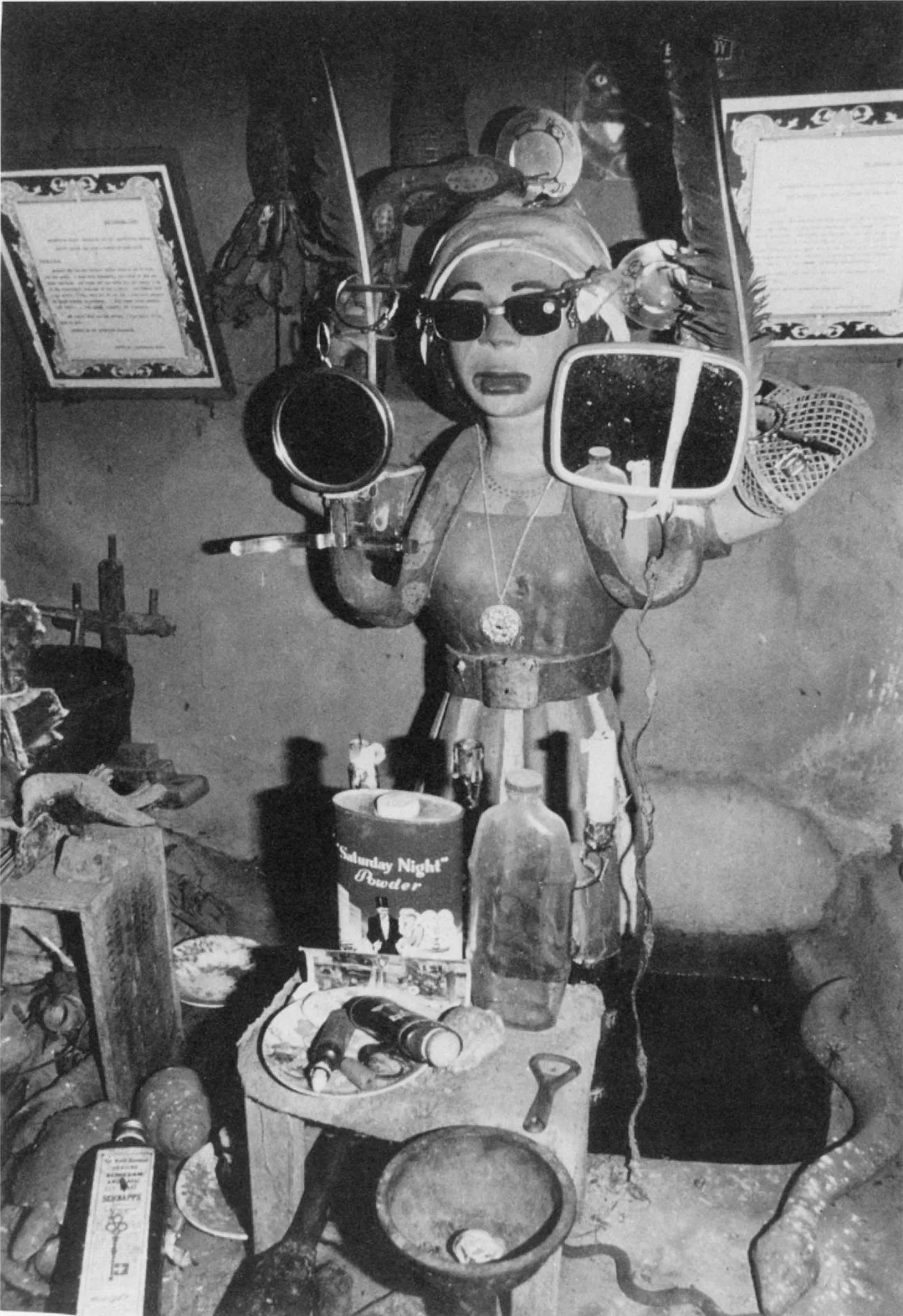 'Mirrors and sunglasses adorn an Igbo shrine sculpture of Mami Wata that stands on Coke bottles in a small pool of water. In front, plates, a bottle, a bottle opener, wooden and metal snakes, talcum powder containers, kola nuts, and a candelabrum clu