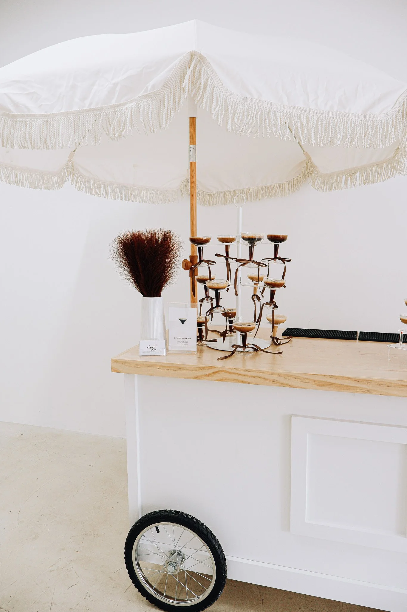 Mobile bar setup with umbrella and cocktail glasses, decorated with limes and plants, on a wooden surface.