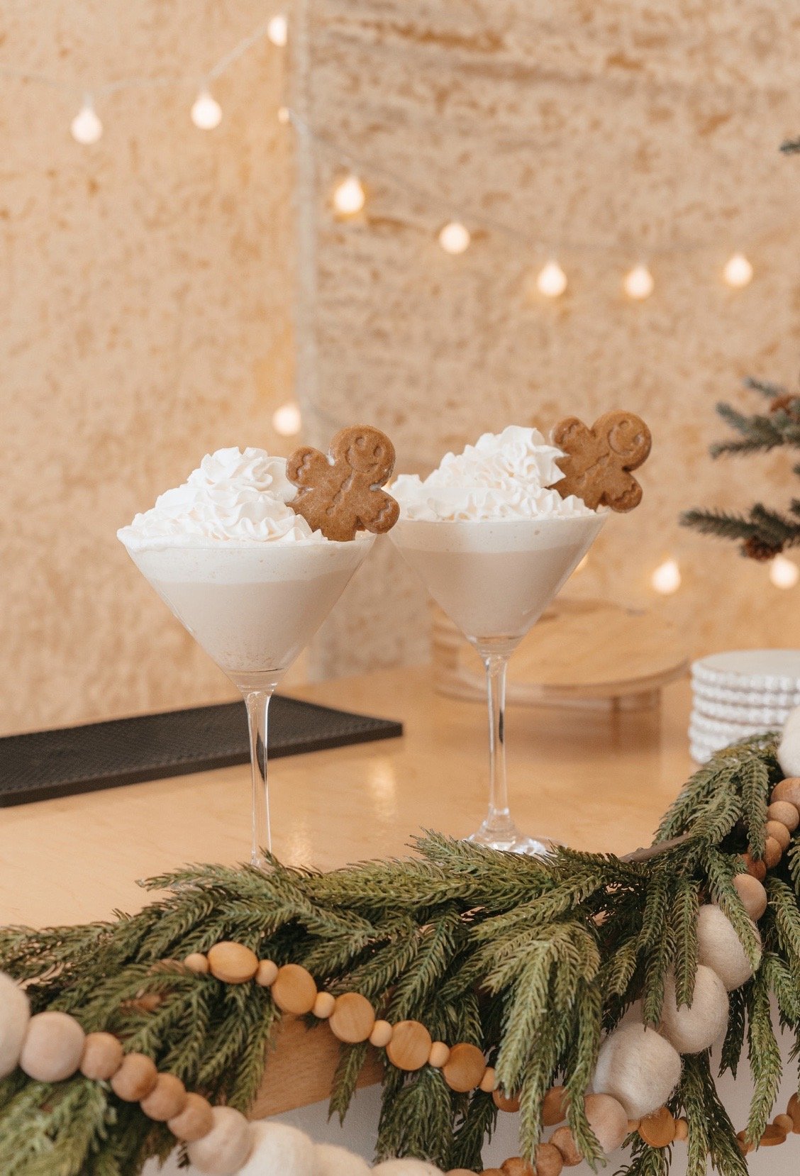 Two festive drinks with whipped cream and gingerbread cookies in martini glasses, on a wooden counter with holiday garland and bokeh lights in the background.