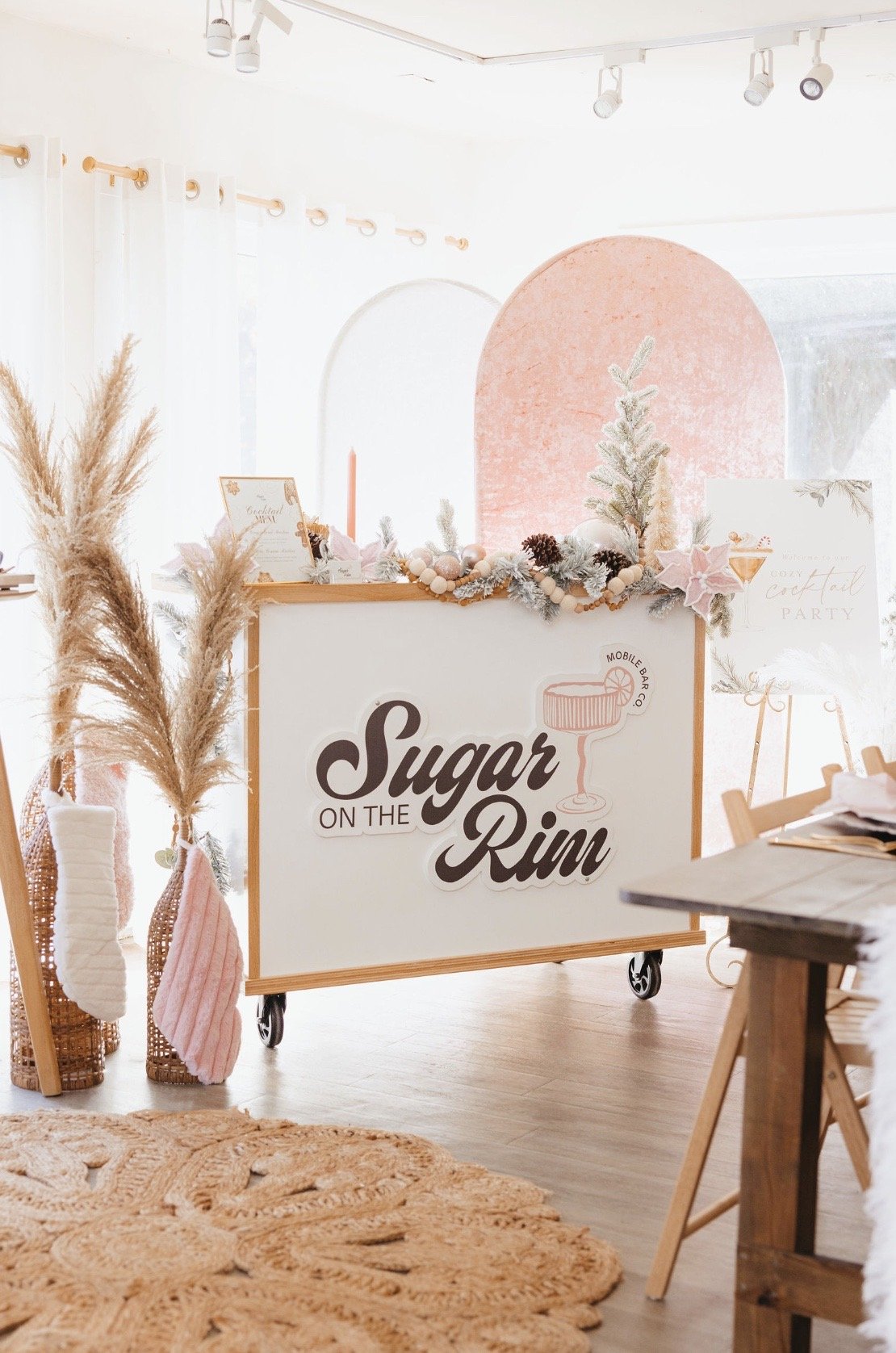 Mobile bar setup with 'Sugar on the Rim' sign and festive winter decorations, including pampas grass, Christmas stockings, and a small artificial tree.