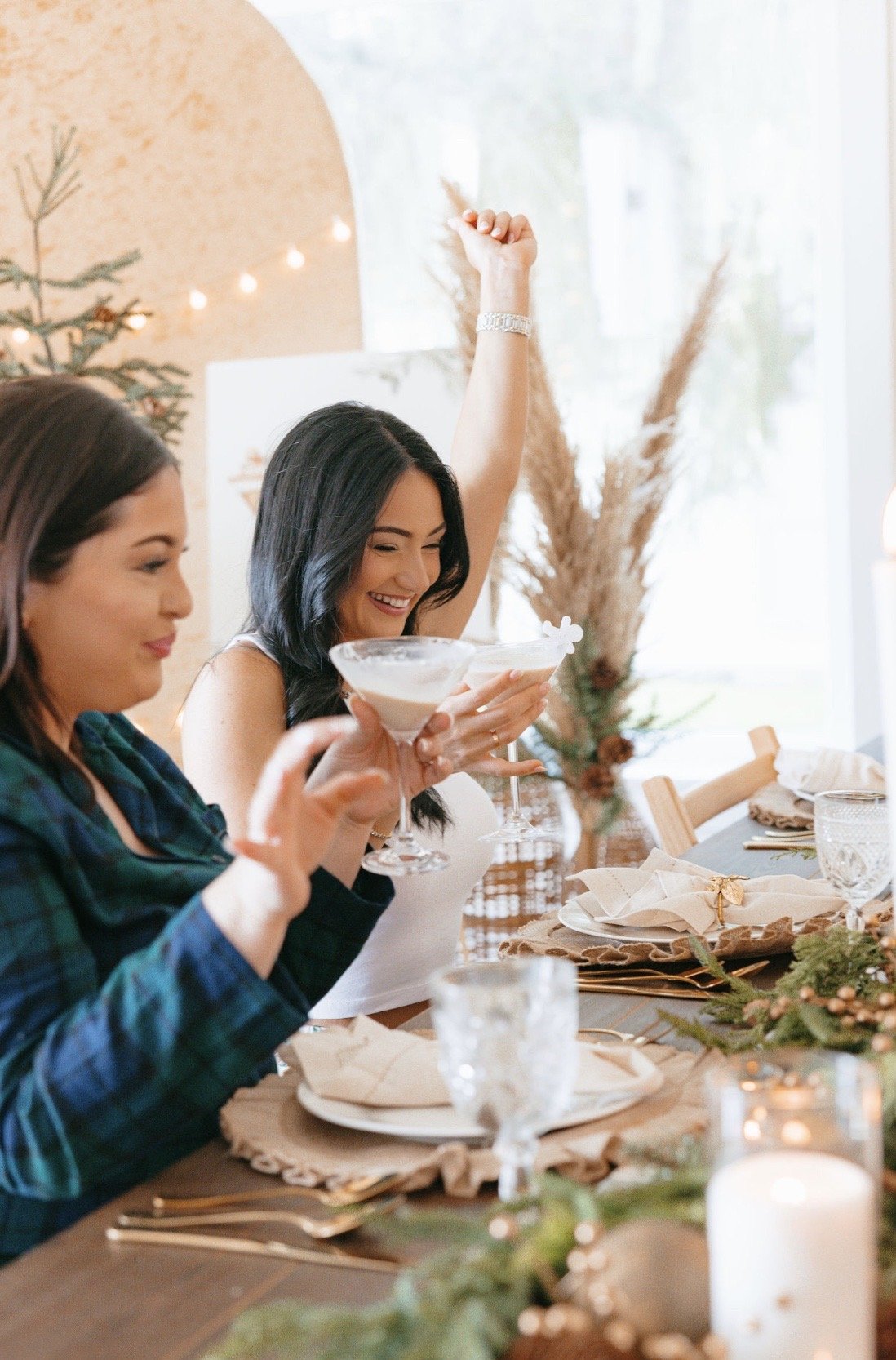 Two women celebrating at a festive dining table, holding cocktails, with holiday decorations and candles.