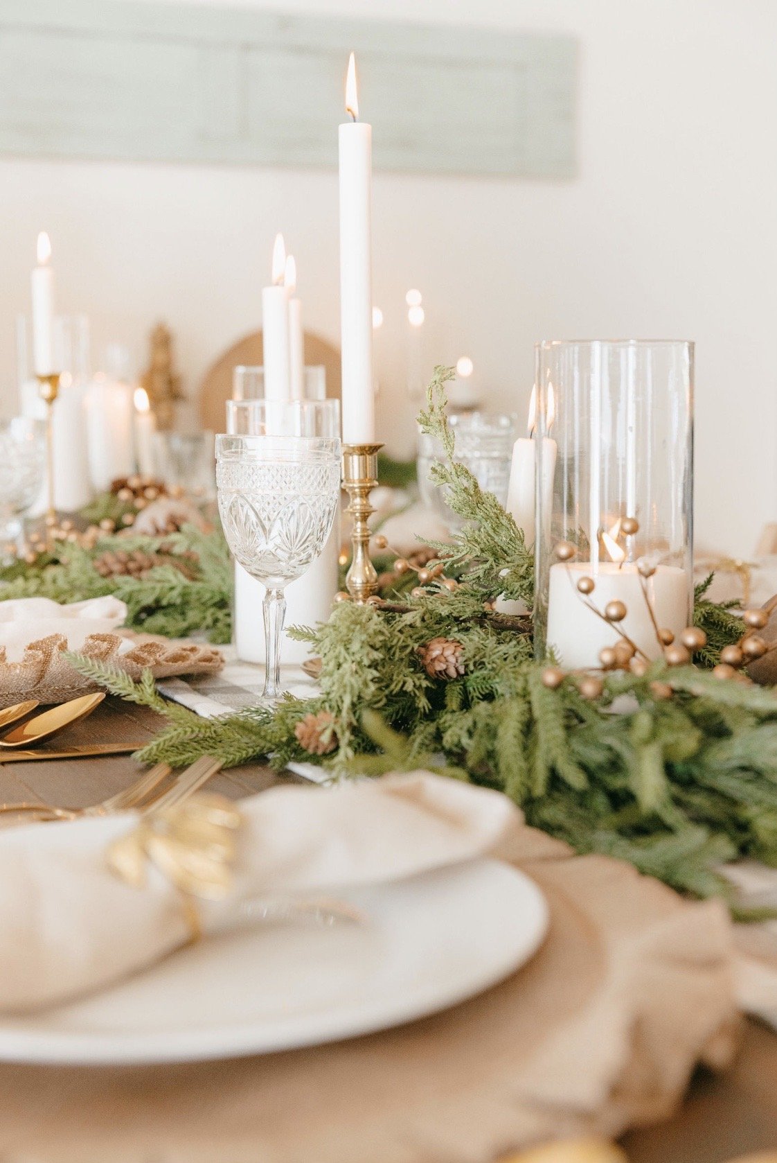 Elegant holiday table setting with candles, greenery, pine cones, glassware, and gold cutlery.