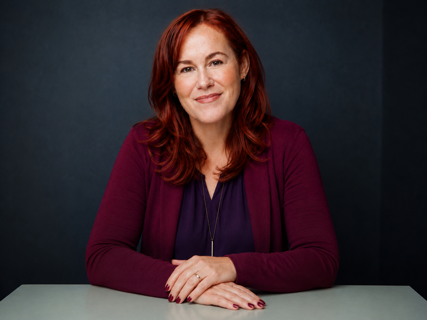 A woman with red hair and a friendly smile, wearing a purple top and maroon cardigan, sitting at a table against a dark background.