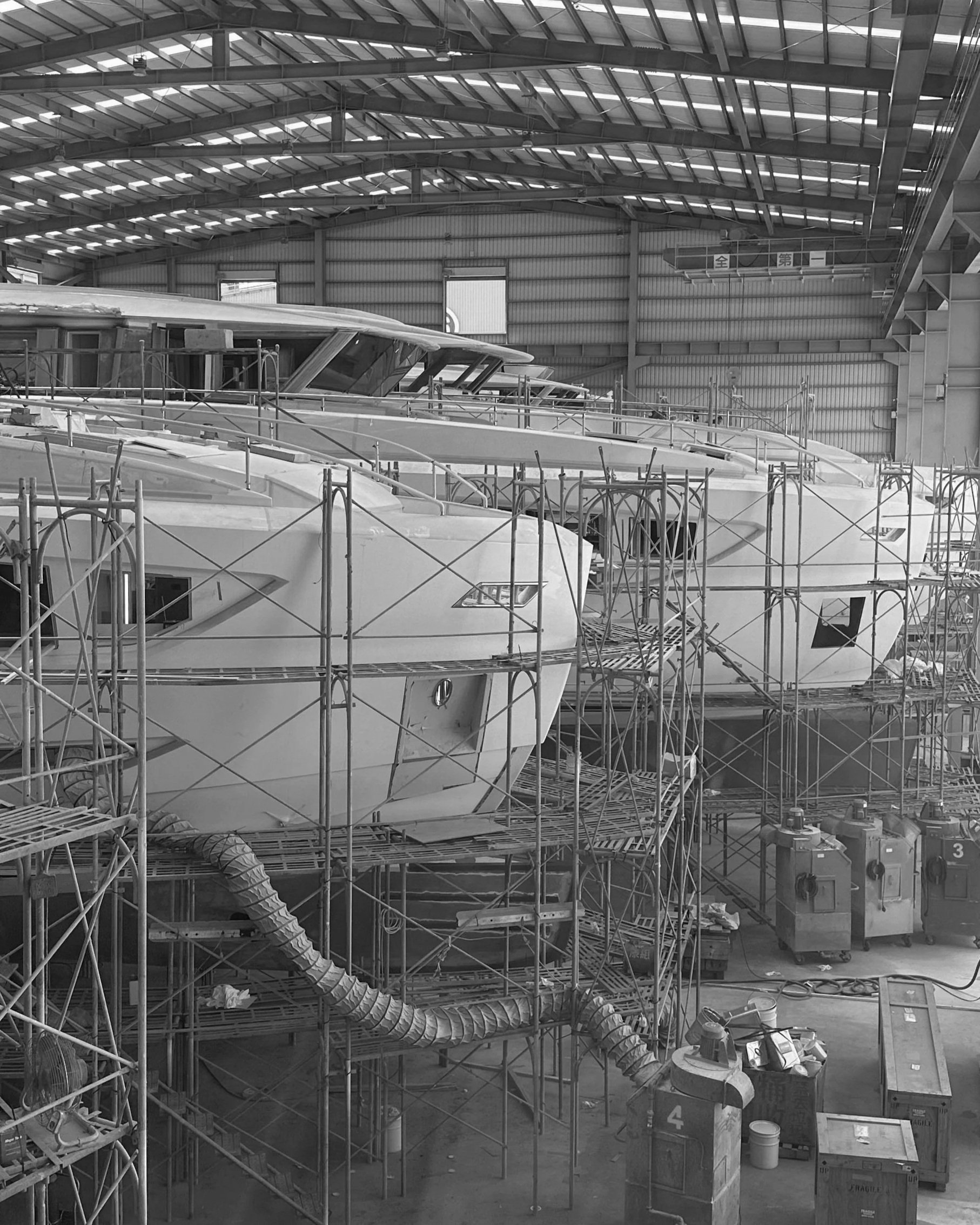 Inside a large industrial warehouse with a steel roof, several yacht hulls are under construction, surrounded by metal scaffolding and super yacht being manufactured