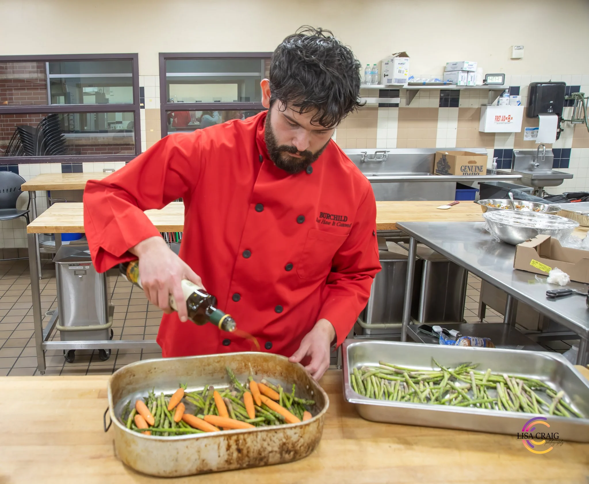 A man with dark hair and beard wearing a red chef's coat preparing vegetables in a kitchen.