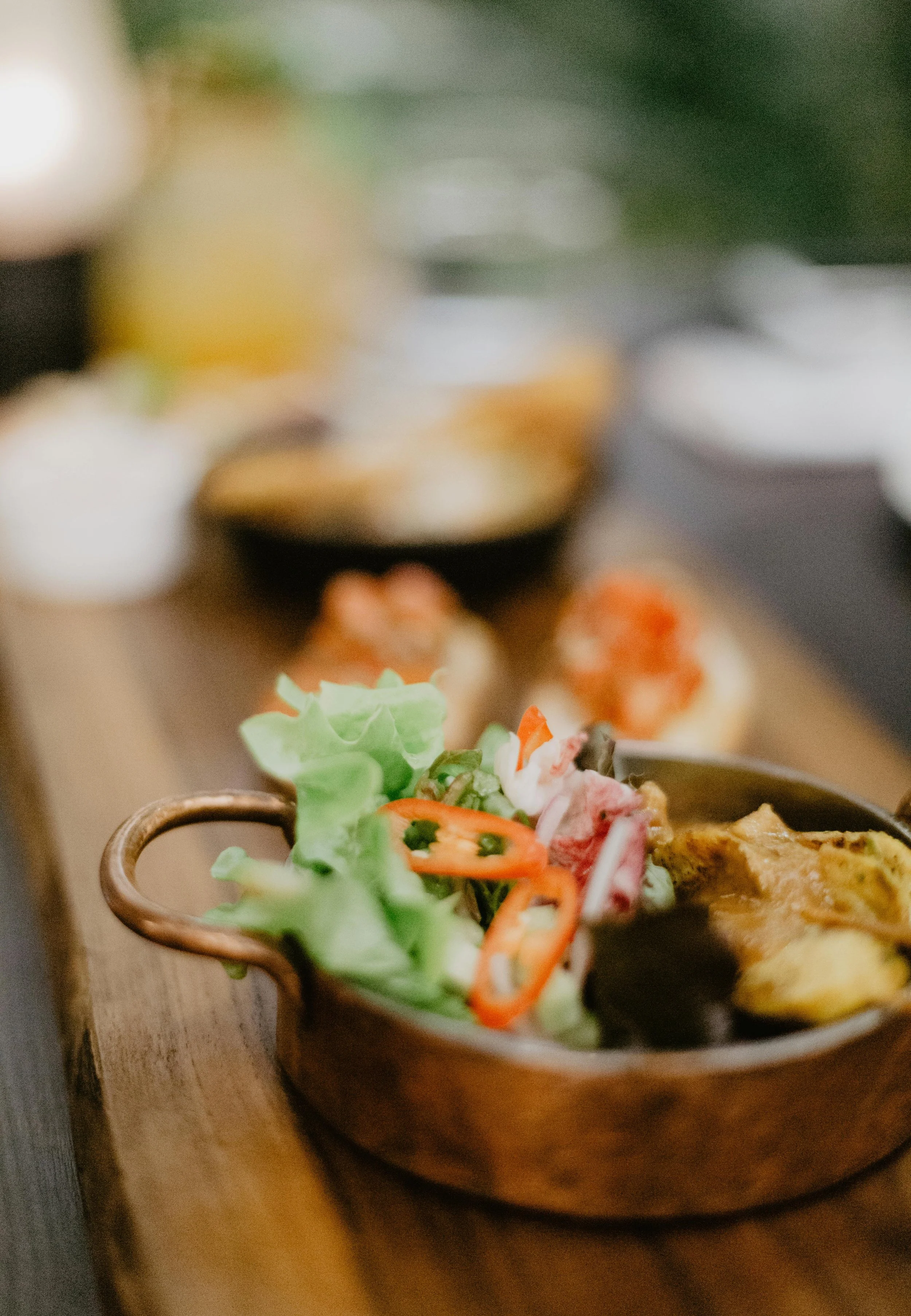 Close-up of a small, brown, ceramic bowl filled with fresh salad, featuring lettuce, tomato slices, and other vegetables, placed on a wooden surface with a blurred background of other dishes.