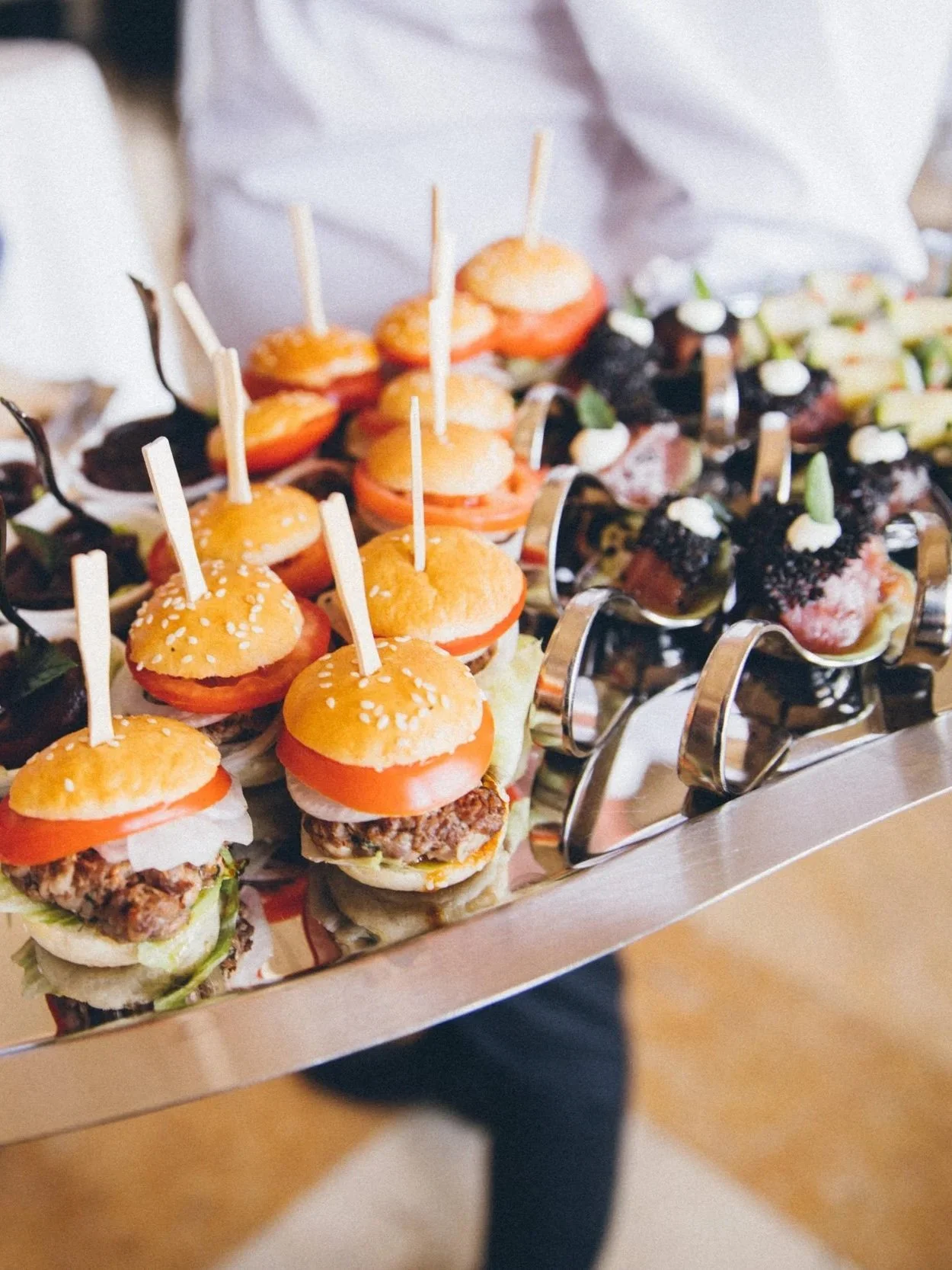 Tray of mini sliders with tomato slices, lettuce, and hamburger patties, and other appetizers with various toppings.
