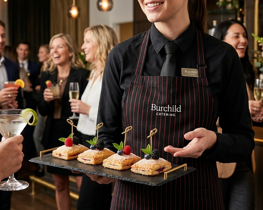 A catering server holds a tray of four mini fruit-filled pastry squares garnished with mint leaves and berries at a party with guests holding drinks
