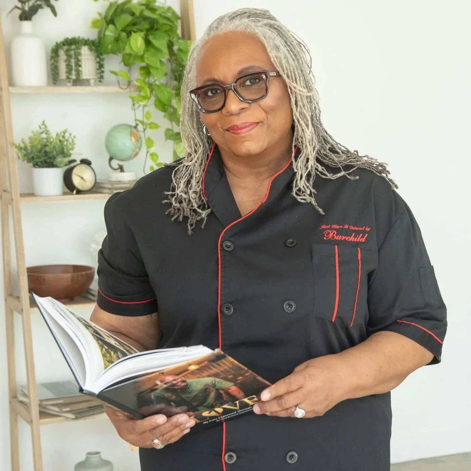 A woman with gray dreadlocks and glasses, wearing a black chef's jacket with red piping, is holding an open magazine or cookbook with food images. She is smiling and standing in a room with shelves, plants, and decorative items in the background.