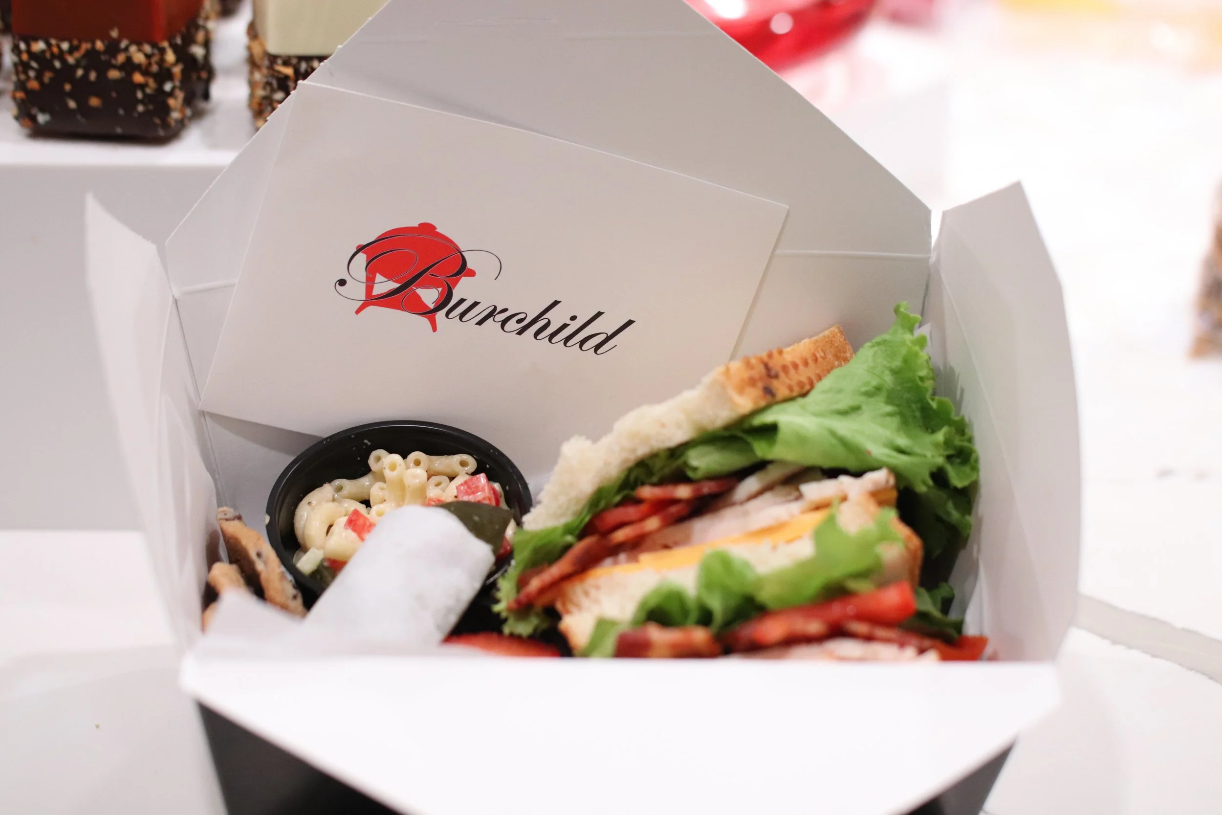 A takeout box with a sandwich, a side of macaroni salad, and a small container of chips, with a logo that reads 'Burchild' on the box.