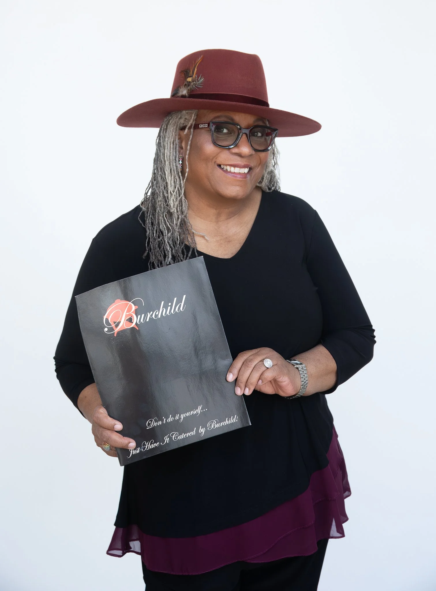 A smiling woman with gray dreadlocks wearing glasses and a red hat with a feather, holding a black sign with pink and white writing in front of a plain white background.