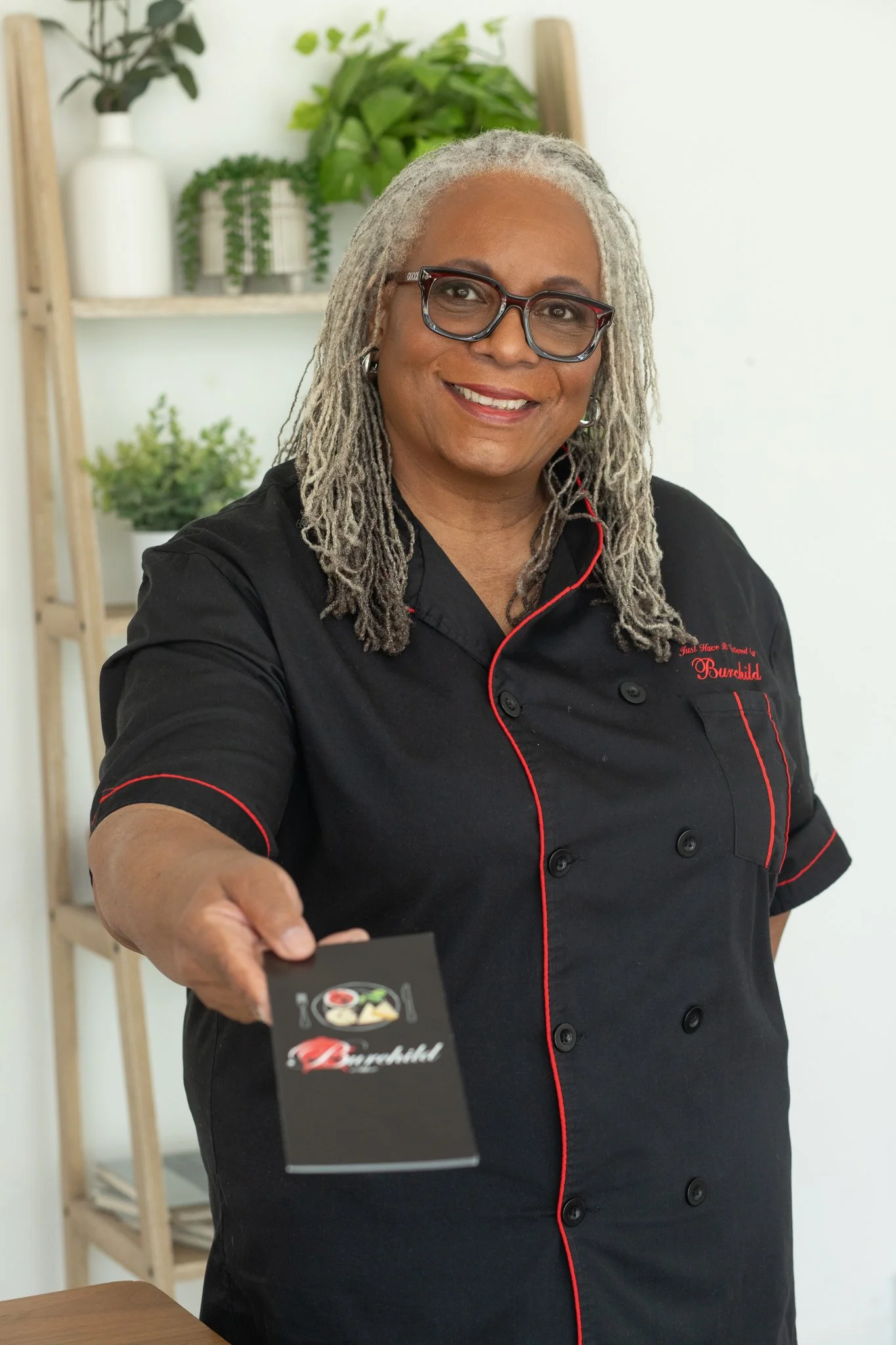 A woman in a black chef's coat with red piping, wearing glasses and earrings, smiling and handing a small black card with a colorful logo to the camera. In the background, there are shelves with potted plants.