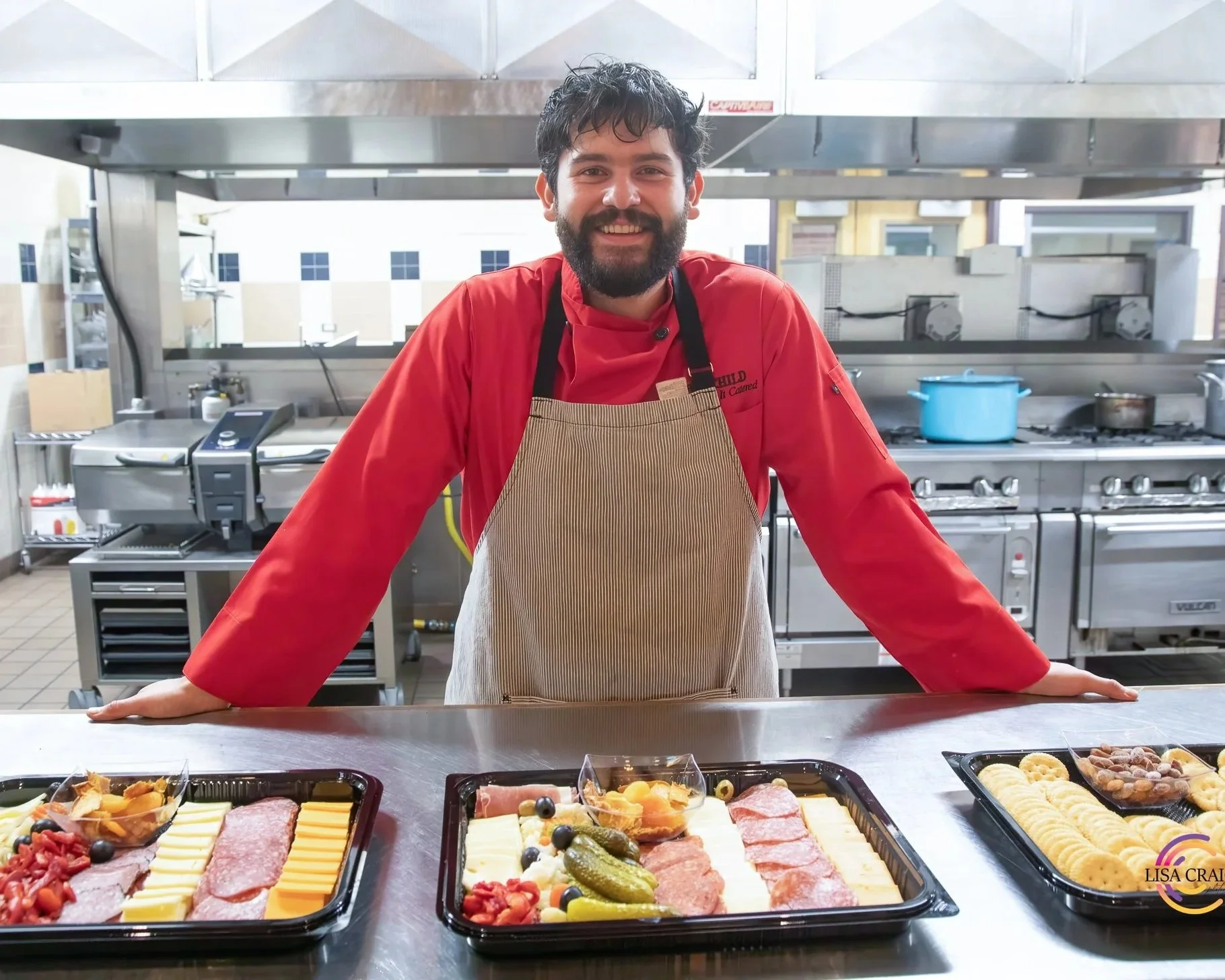 A man with a beard and dark hair wearing a red chef's jacket and beige apron, smiling behind a kitchen counter with trays of assorted foods including meats, cheese, and vegetables. The kitchen has stainless steel appliances and a window with buildings visible outside.