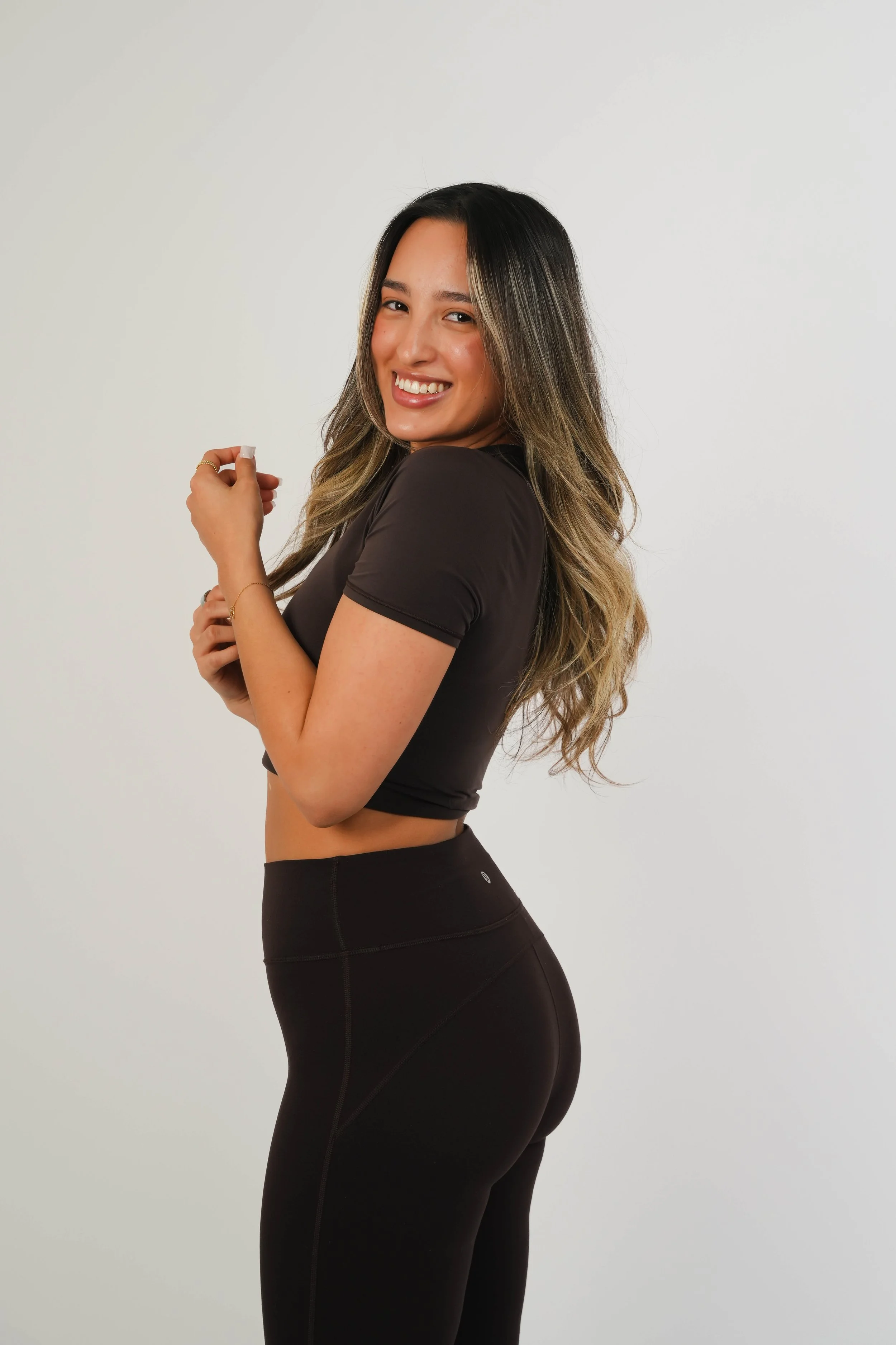 A young woman with long, wavy hair smiling and looking at the camera, dressed in a black athletic top and leggings, standing against a plain white background.