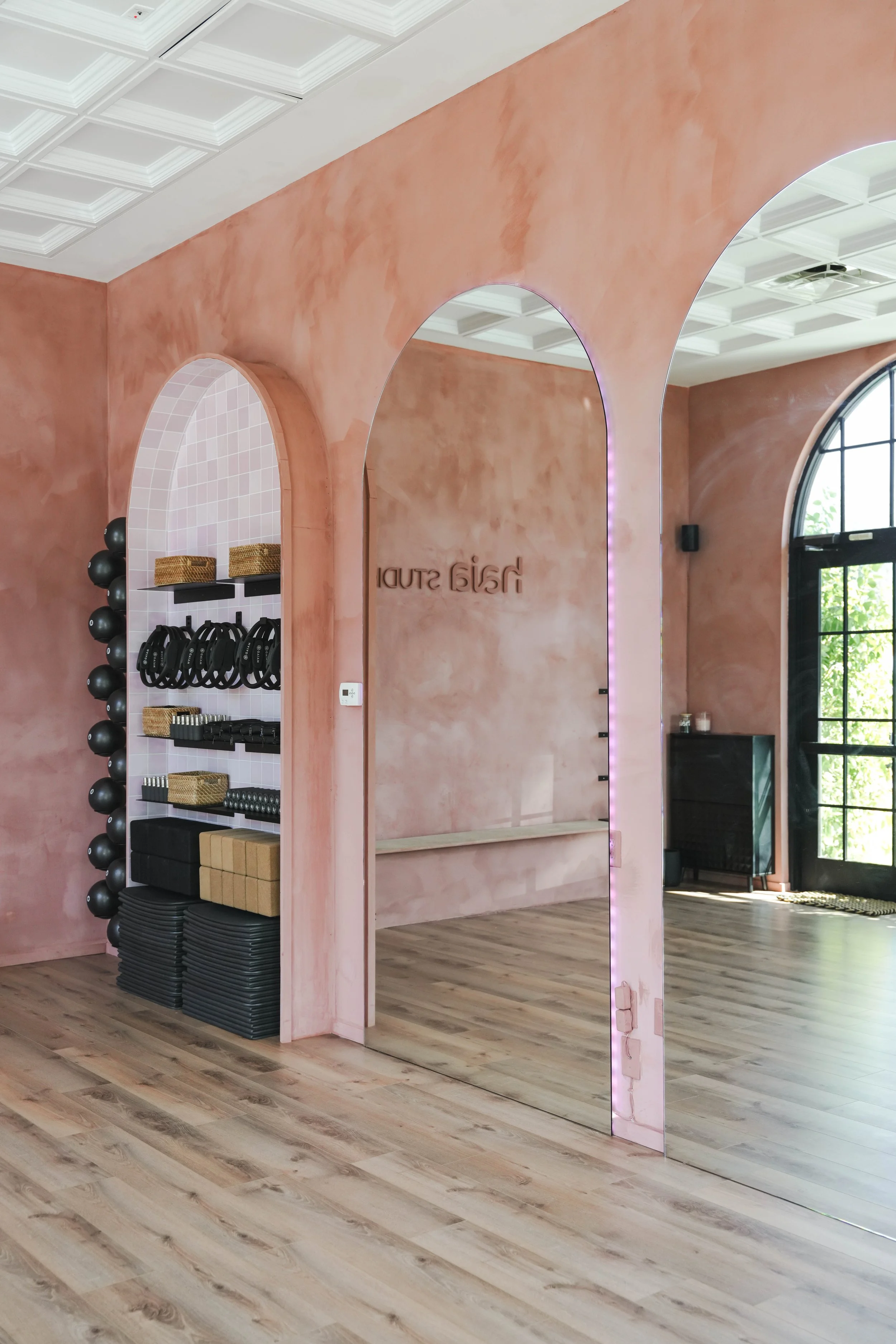 Interior of a fitness studio with pink walls, arched mirrors, and workout equipment including black medicine balls, weights, and mats. Natural light from large windows illuminates the space.