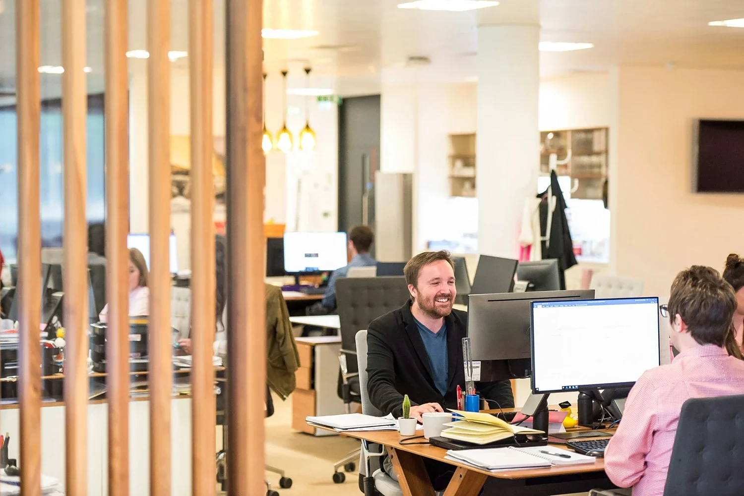 Office workspace with employees working at desks, some focused on computers, and one man smiling and engaging in conversation, surrounded by office furniture and decor.