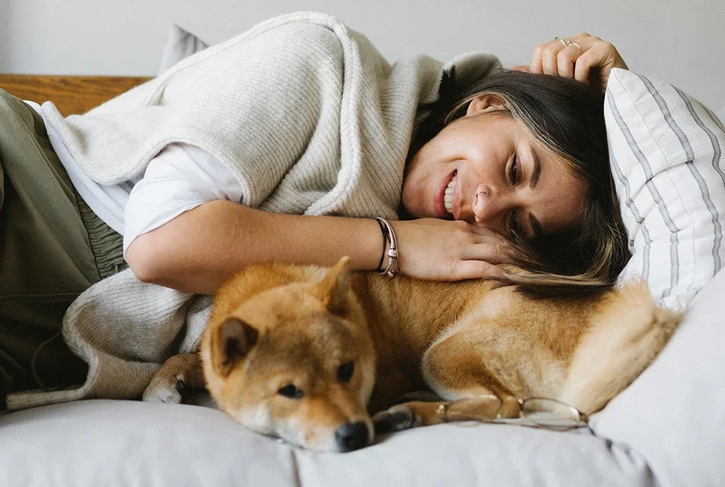 Woman with brown hair laying down cuddling with her dog. The woman is smiling and comfortable.