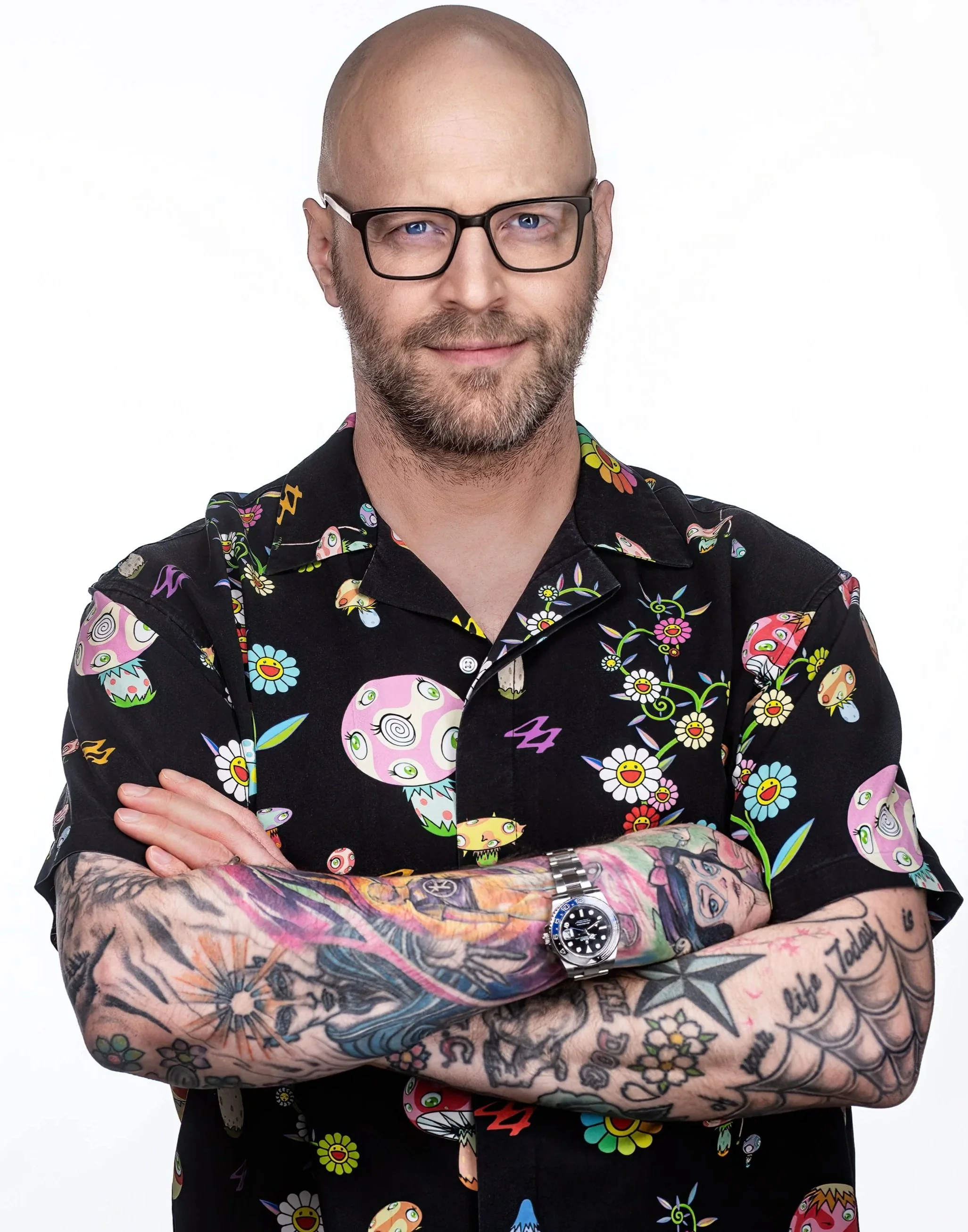 A man with glasses, a beard, and tattoos on his arms, wearing a colorful, patterned short-sleeve shirt, crossing his arms and smiling against a white background.