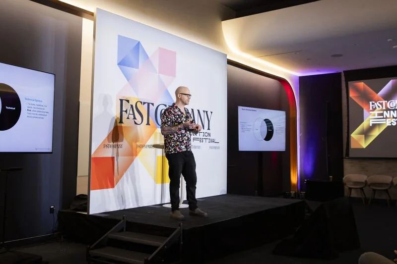 A man in a colorful floral shirt and black pants speaking on stage at the Fast Company Innovation Festival, with multiple screens displaying presentation slides and a large backdrop with the festival's logo.
