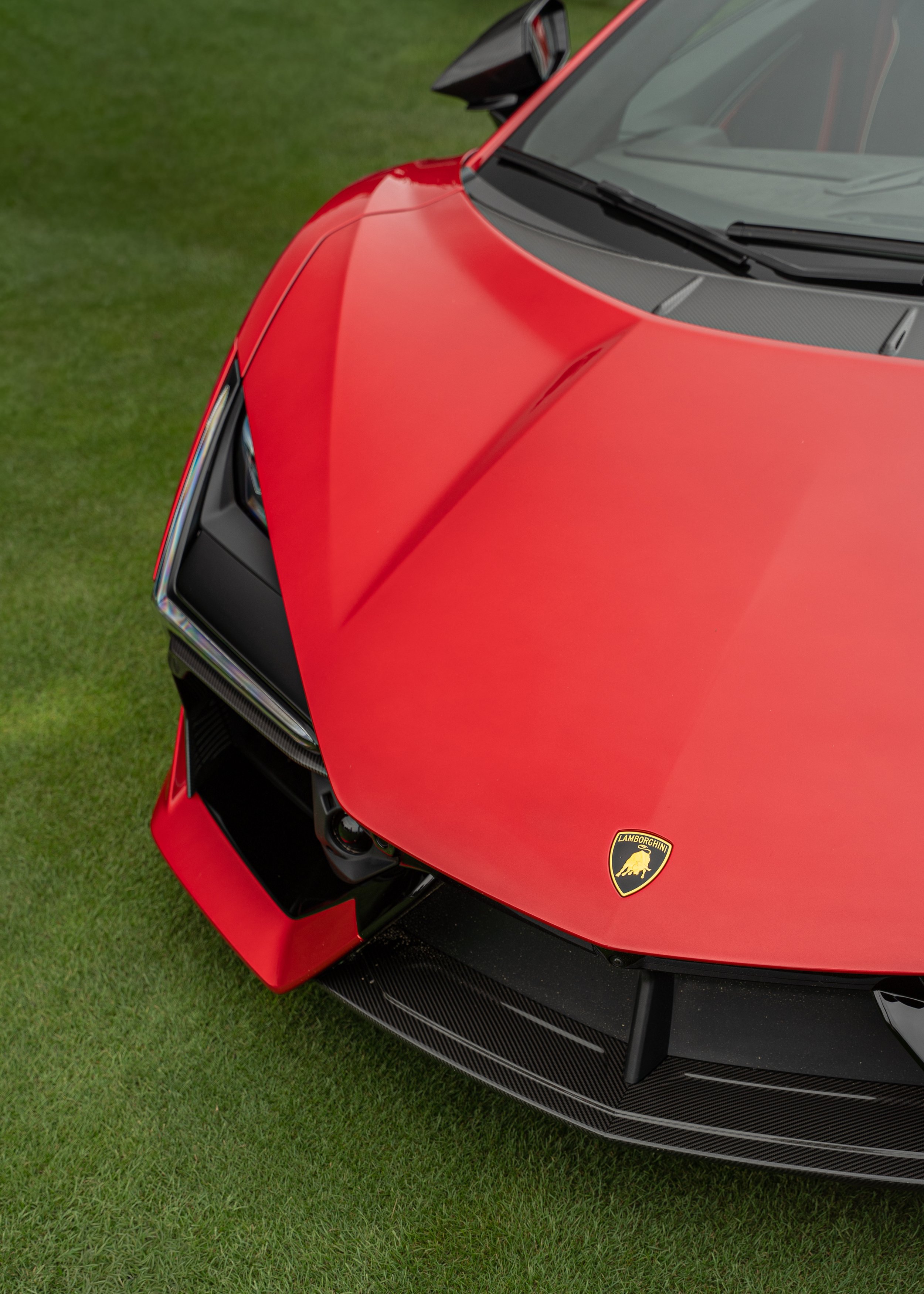 Close-up of the front of a red Lamborghini car on grass, showing the Lamborghini emblem and part of the headlight and black front splitter.