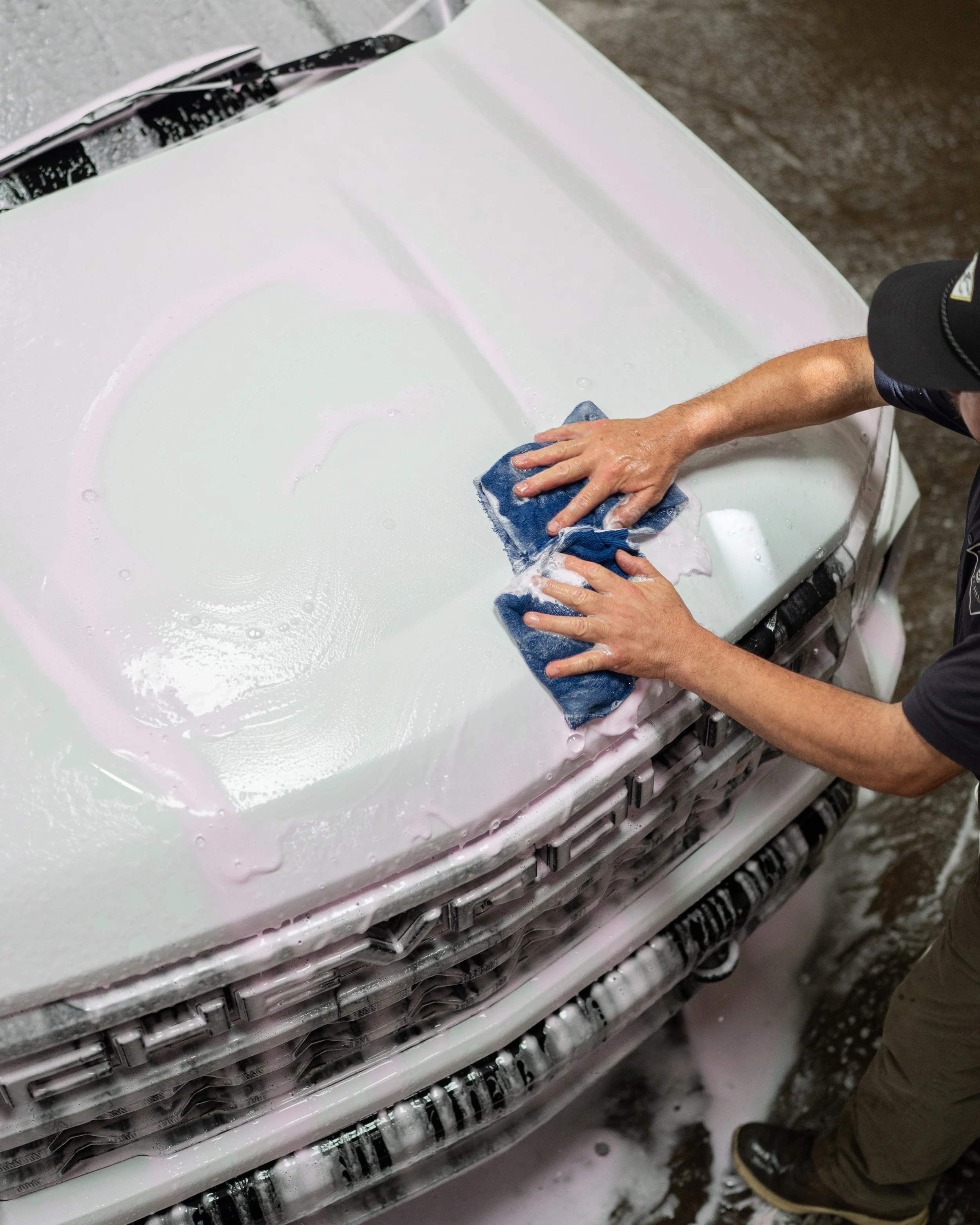Person cleaning the front of a white vehicle using a blue cloth, with soap suds and bubbles visible on the surface. The vehicle parts and a person wearing khaki pants and a black cap are partially visible.