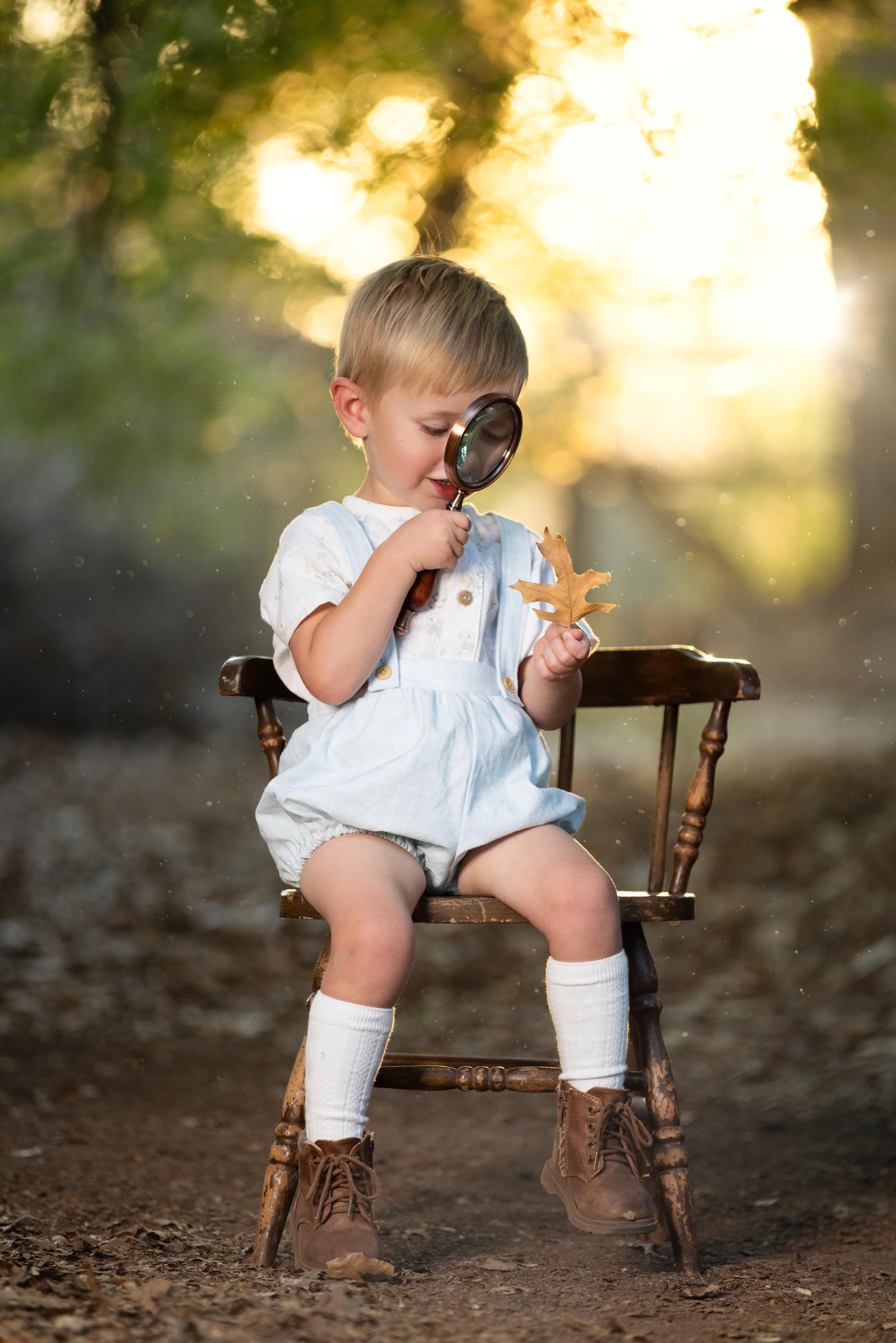Whimsical Children's portrait of a toddler sitting on a vintage chair looking at a leaf through a magnify glass at the tonaquint nature center in saint George Utah.