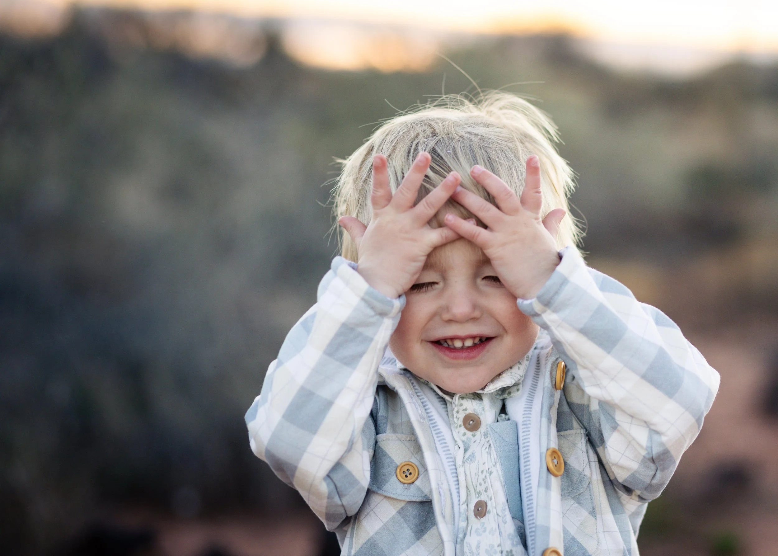 A young child with blonde hair smiling and covering their face with both hands. The child is wearing a plaid jacket with buttons and a shirt underneath. Lava rock background at sunset in southern Utah.