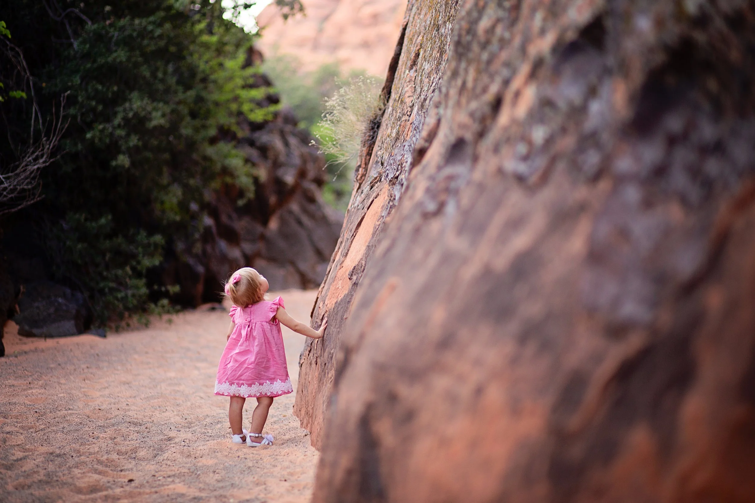 Toddler wearing pink dress walking in sand look up at large red sandstone rock