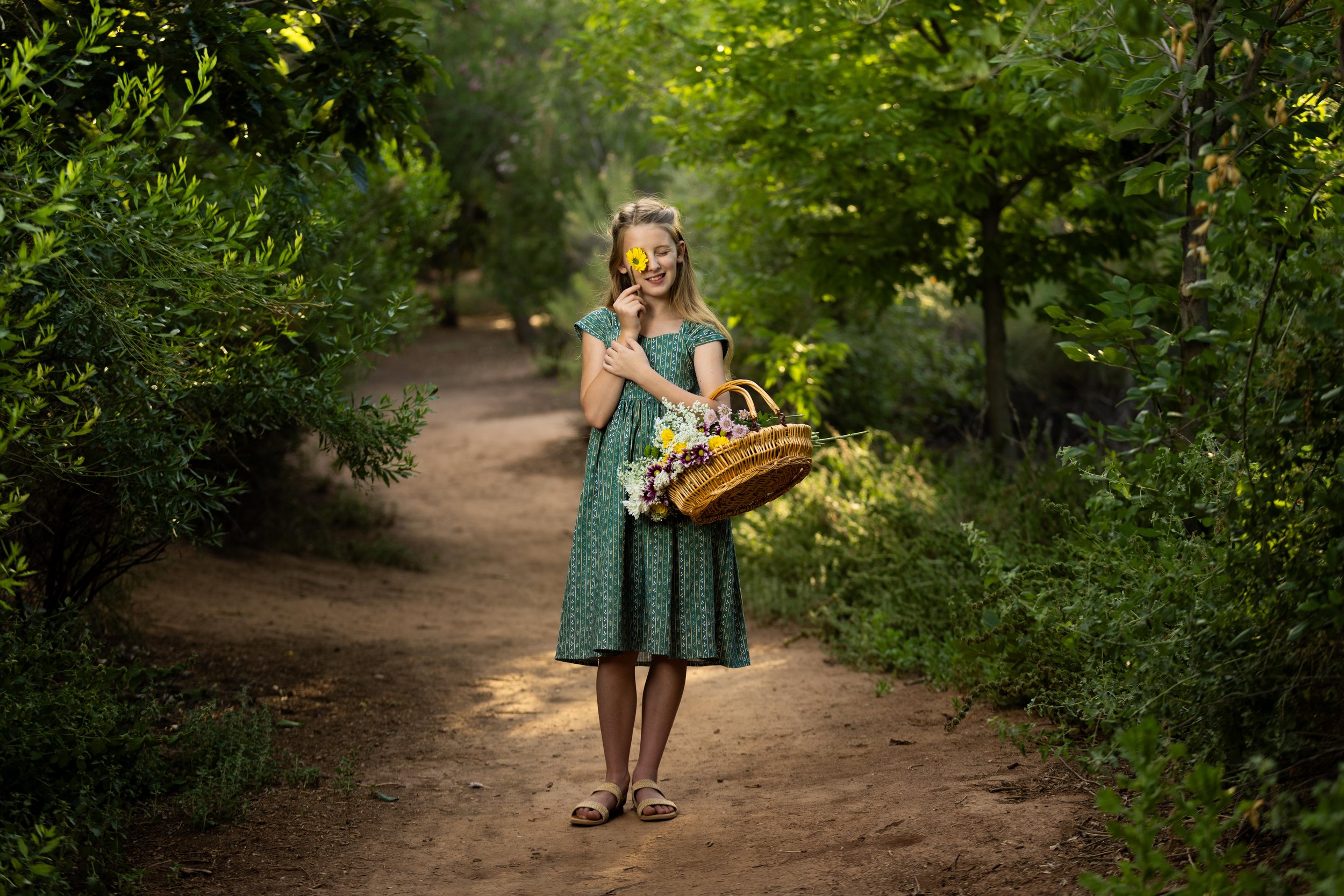 A young girl with long blonde hair standing on a dirt path surrounded by lush green trees, holding a yellow flower in front of her face and carrying a basket filled with colorful flowers. Tonaquiant nature center in the summer.