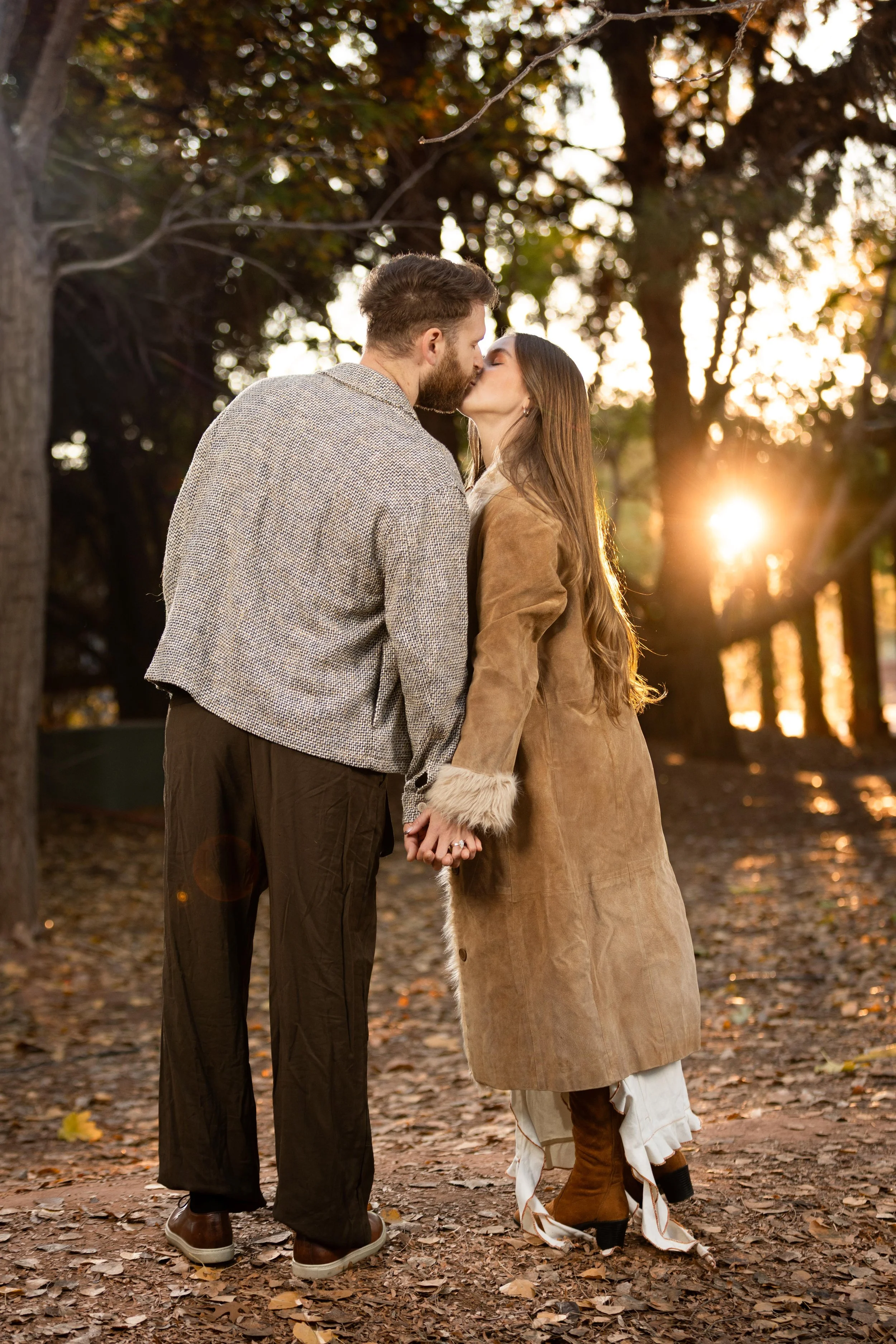 engaged couple kissing under the autumn trees with the golden light of the sun behind them.