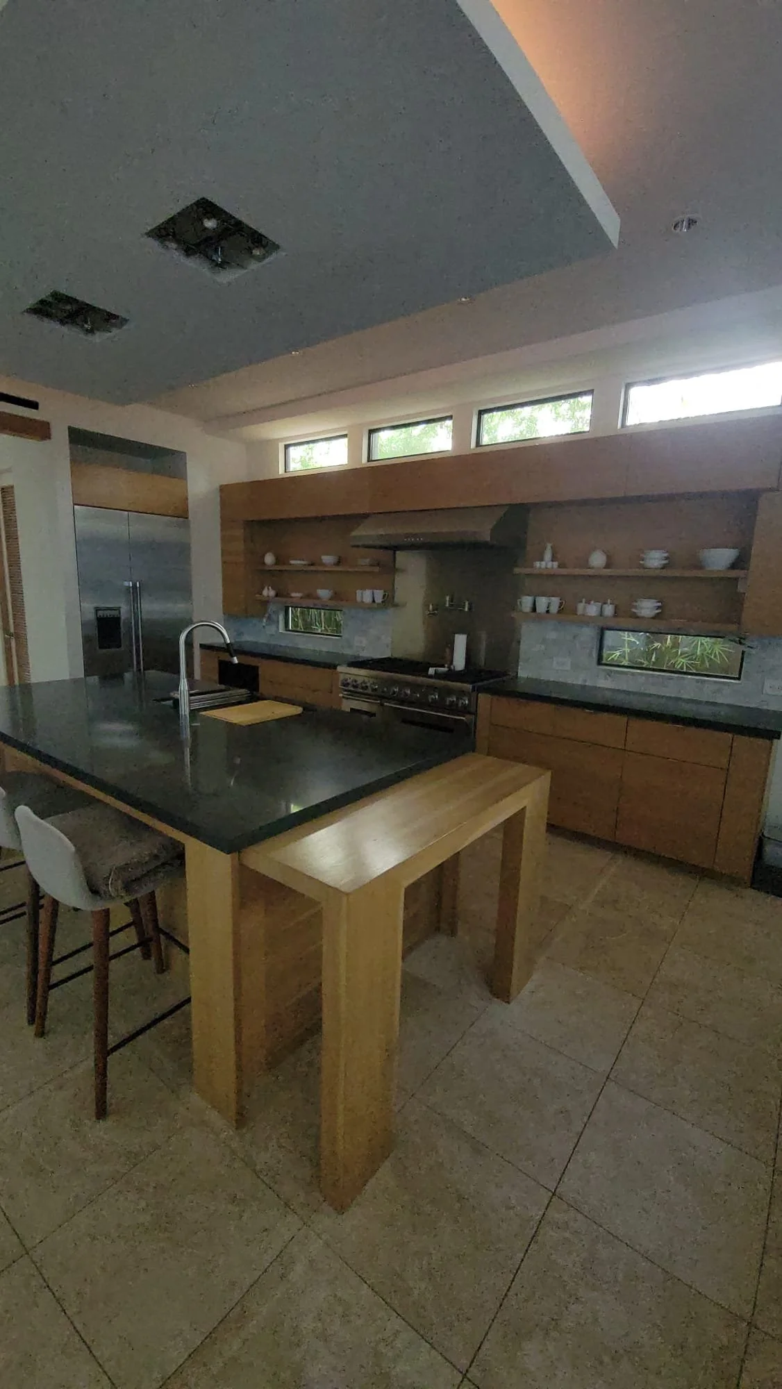 Modern kitchen with large island, bar stools, wooden cabinetry, stainless steel appliances, and open shelving, under a high ceiling with natural light.
