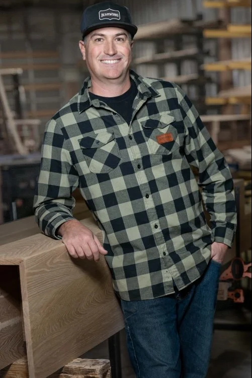 A smiling man in a black and green plaid shirt and a black baseball cap standing in a woodworking shop.