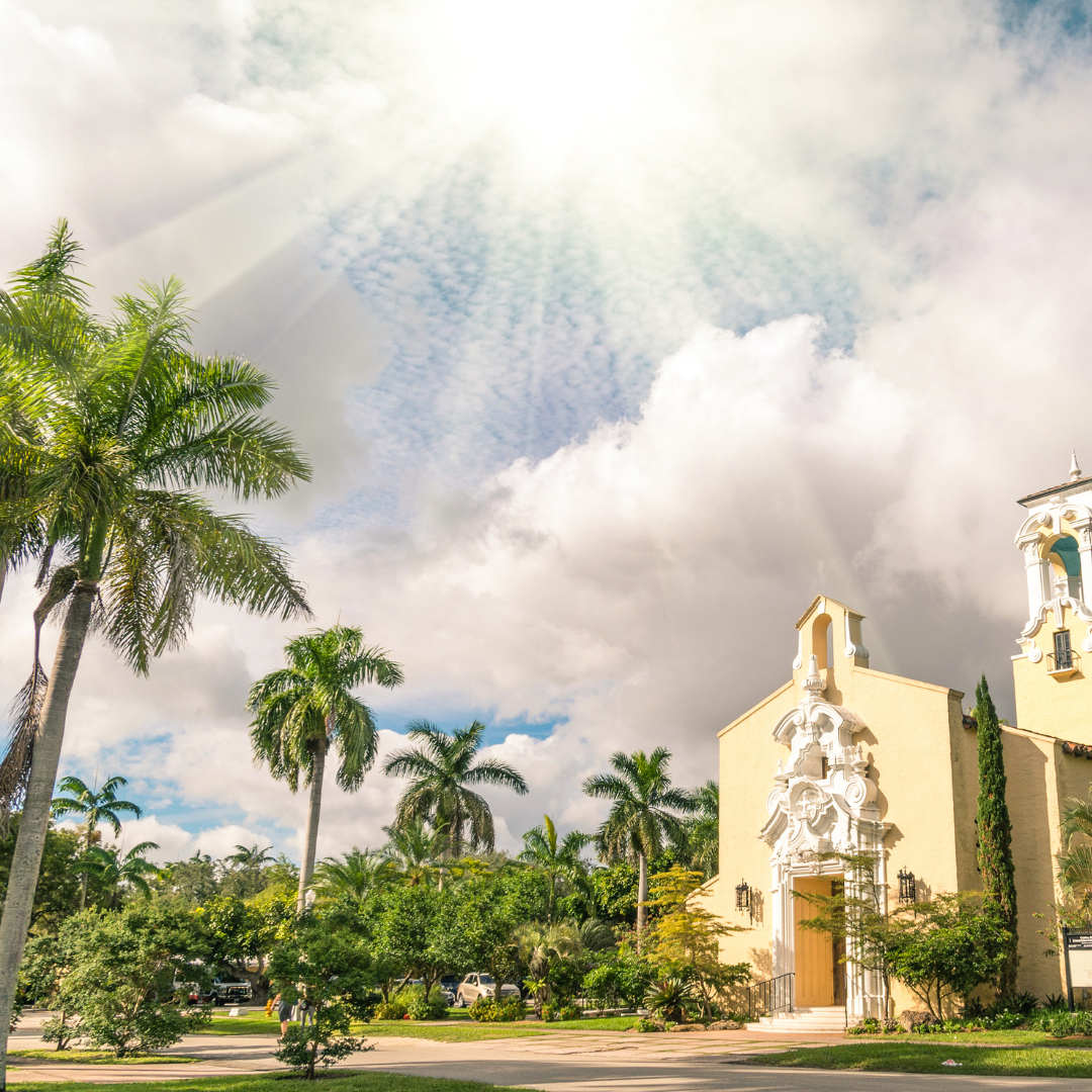 The sun shines down on the Coral Gables Congregational United Church of Christ. There are some clouds in the sky and some palm trees in the foreground.