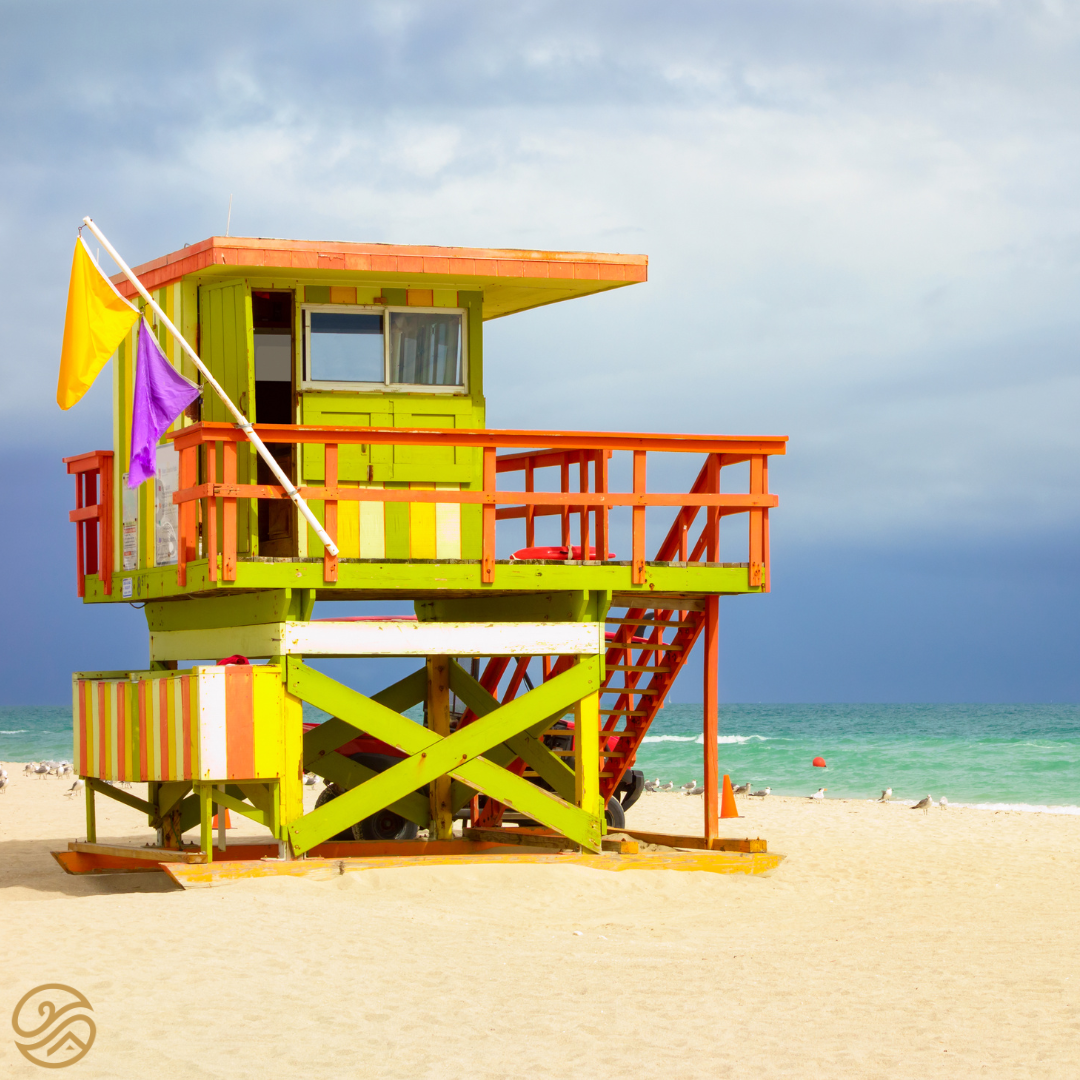 A bright, neon lifeguard tower is in the foreground of the beach, with a moody sky and ocean in the background.