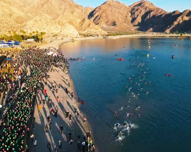 Overhead view of a crowded beach event with a long red carpet, palm trees, tents, and a pier extending into the ocean near a city shoreline.