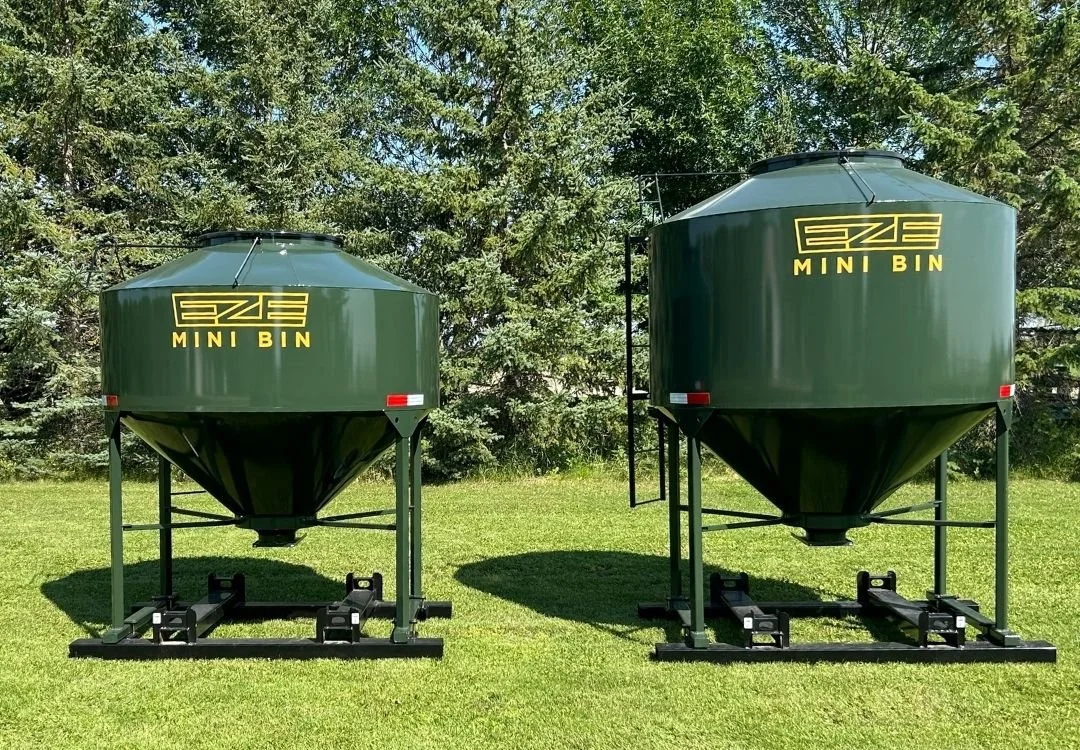 Two EZE Mini Bin,  green feed bins sitting side by side on a green lawn with evergreen trees in the background.