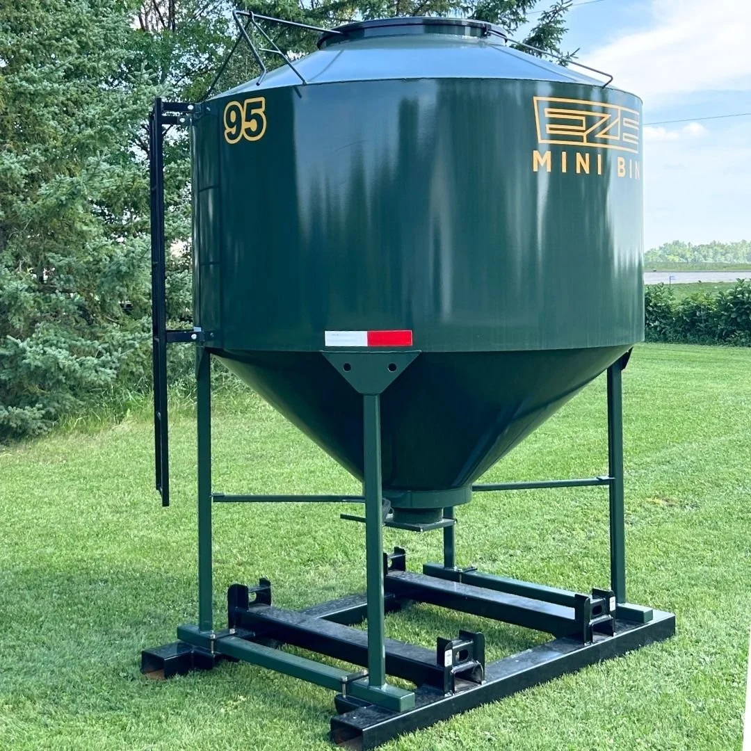 Side view of a green feed storage bin, 95 bushel EZE Mini-Bin  sitting on lush green grass with trees in the background