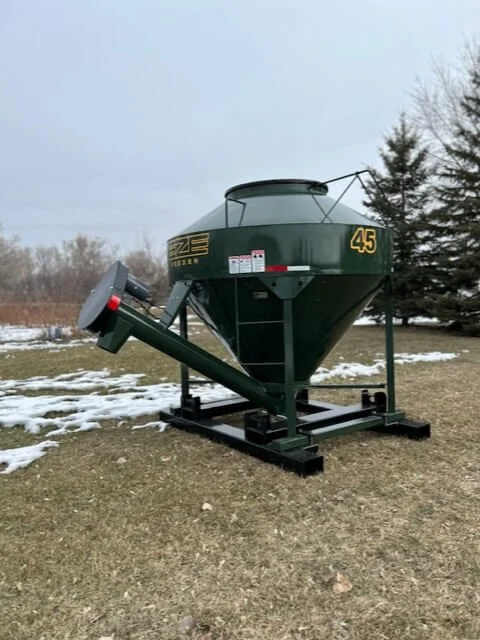 Outdoor scene with a large green grain hopper with the number 45 on it, placed on a black metal stand on a grassy field with patches of snow, and a few trees in the background.