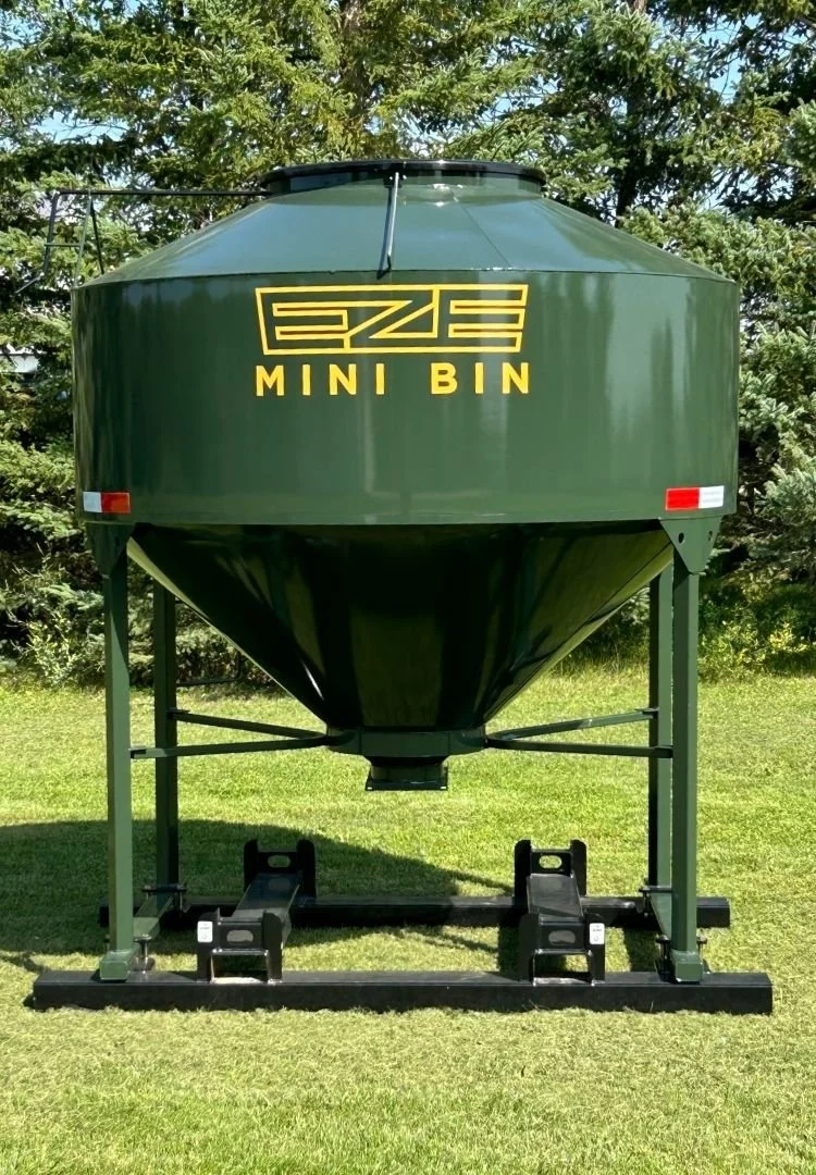 Front view of a dark green 70 Bushel Mini-Bin feed storage hopper sitting on green grass with trees behind it.