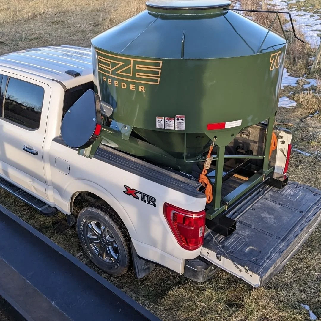A White Ford pickup truck in a field with a green EZE Feeder cattle feeder tied down on the back with orange tie downs.