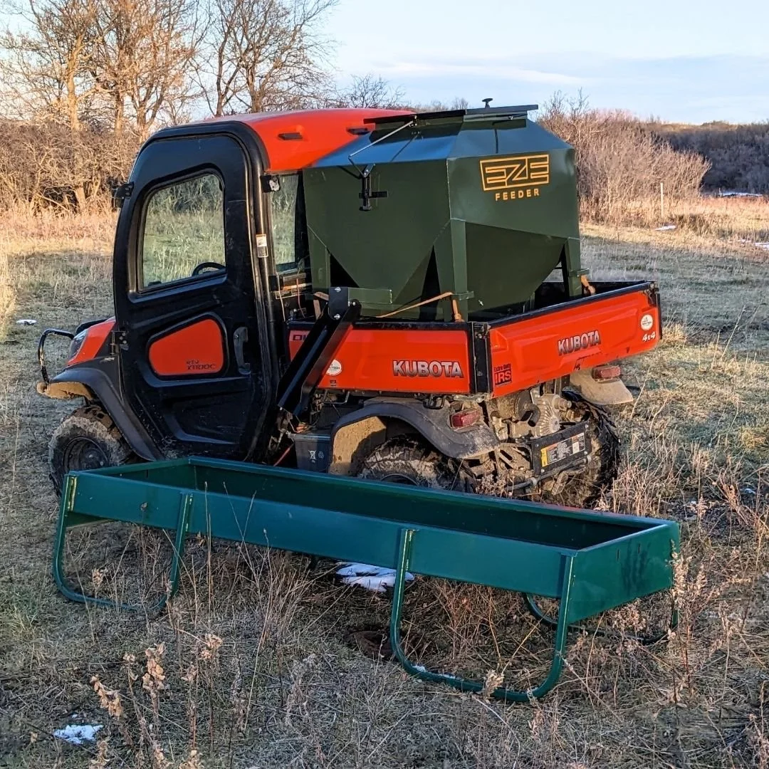 A green utv15 ivestock feeder with a black spout in the back of a koboda UTV located in a feild with dry grass and bare trees in the background