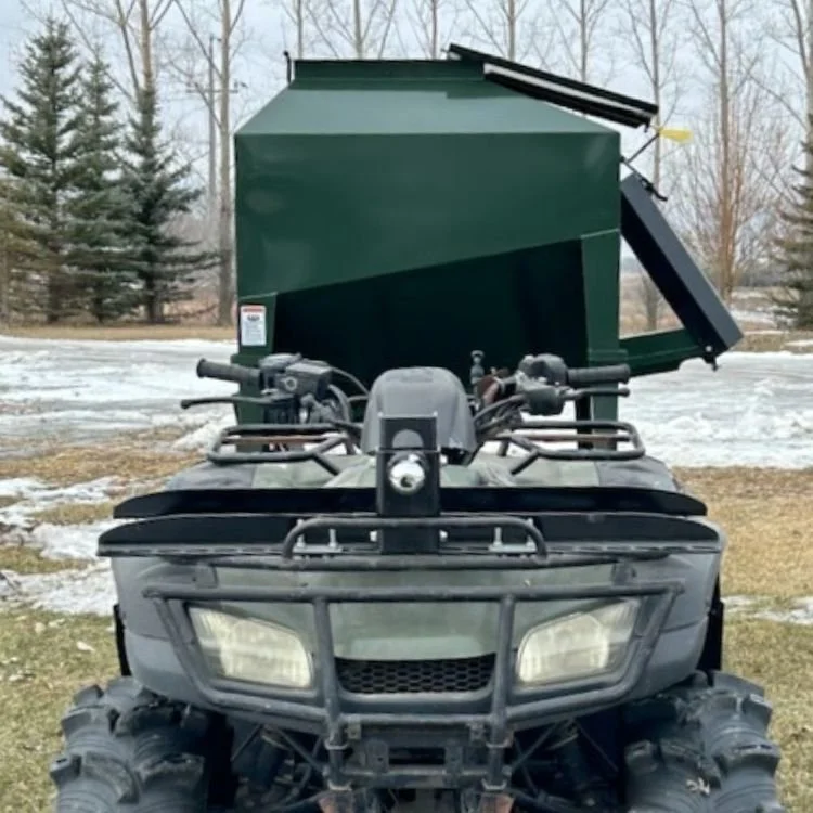 Front view of an all-terrain vehicle (ATV) with a black and gray body, hooked up to a tow behind portable cattle feeder, there is snow in the background and evergreen trees