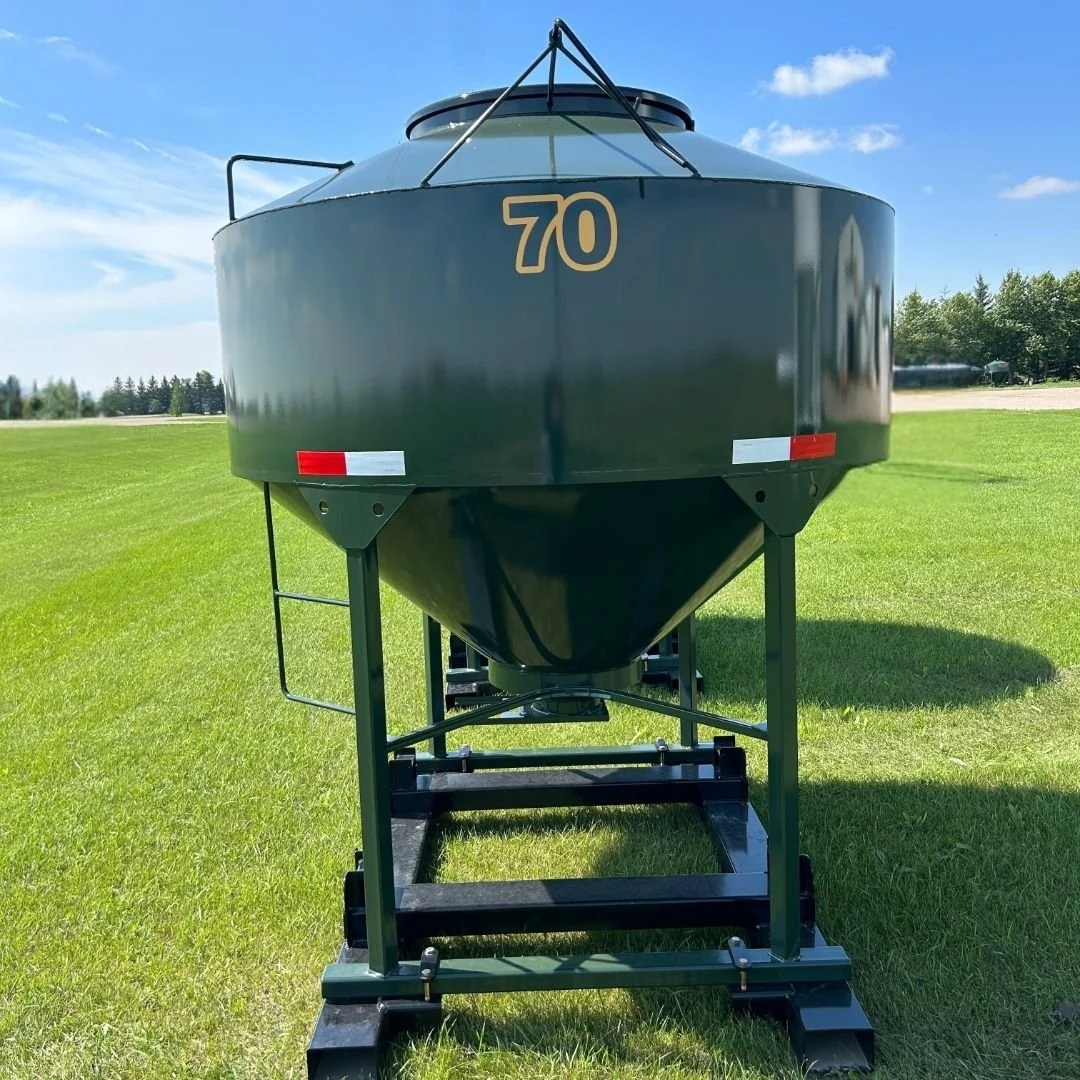 Back view of an EZE Mini Bin grain storage tank with 70  logo for 70 bushel, a black metal frame with fork pockets.  Sitting on green grass with blue skies, a few white fluffy clouds and evergreen tress in the distance.