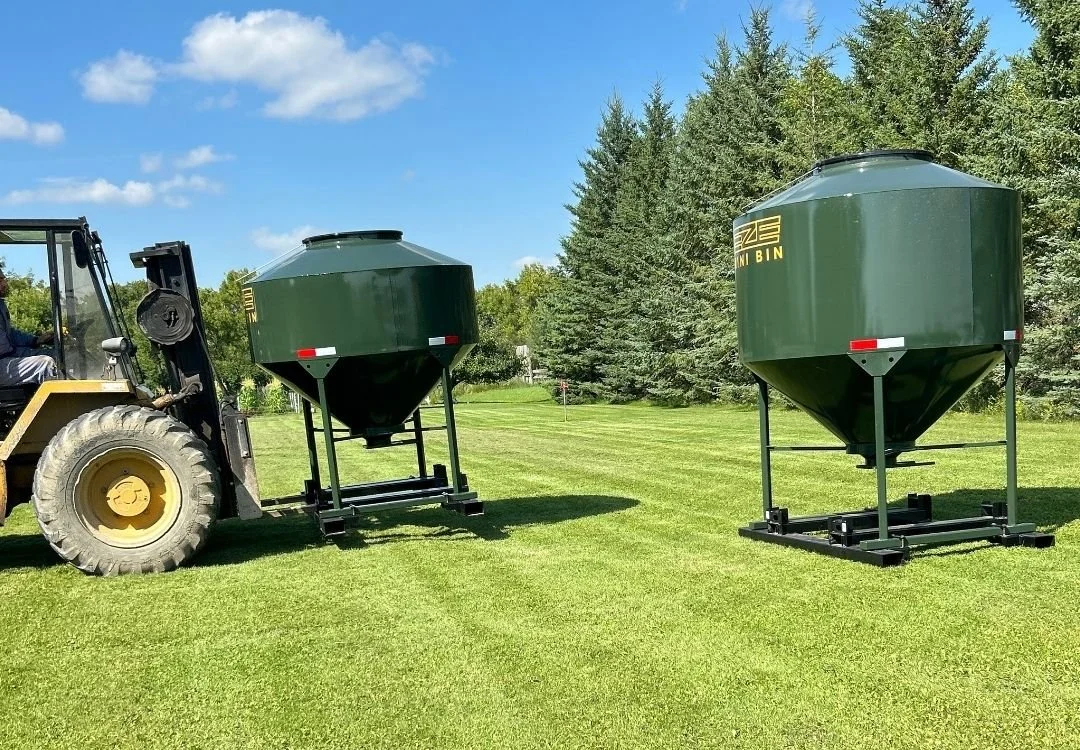 A yellow fork lift moving a green 70 bushel EZE Mini-Bin, grain storage tank, with a larger tank close by , green grass , blue skies and healthy evergreen trees in a row.
