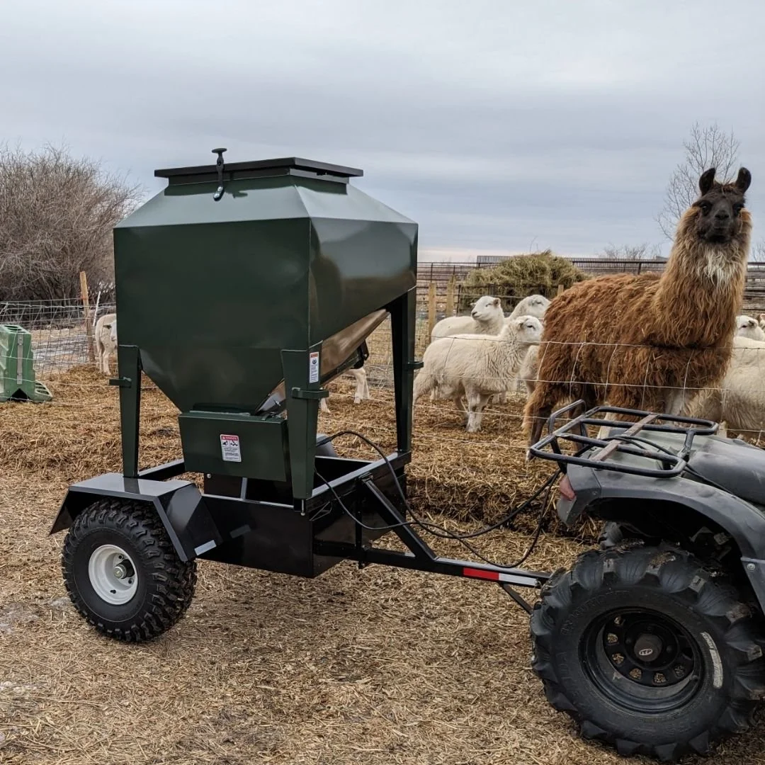 ATV feeder towed behind a quad, feeding white sheep and a  brown llama inside a farm pen with dark skies in the background.